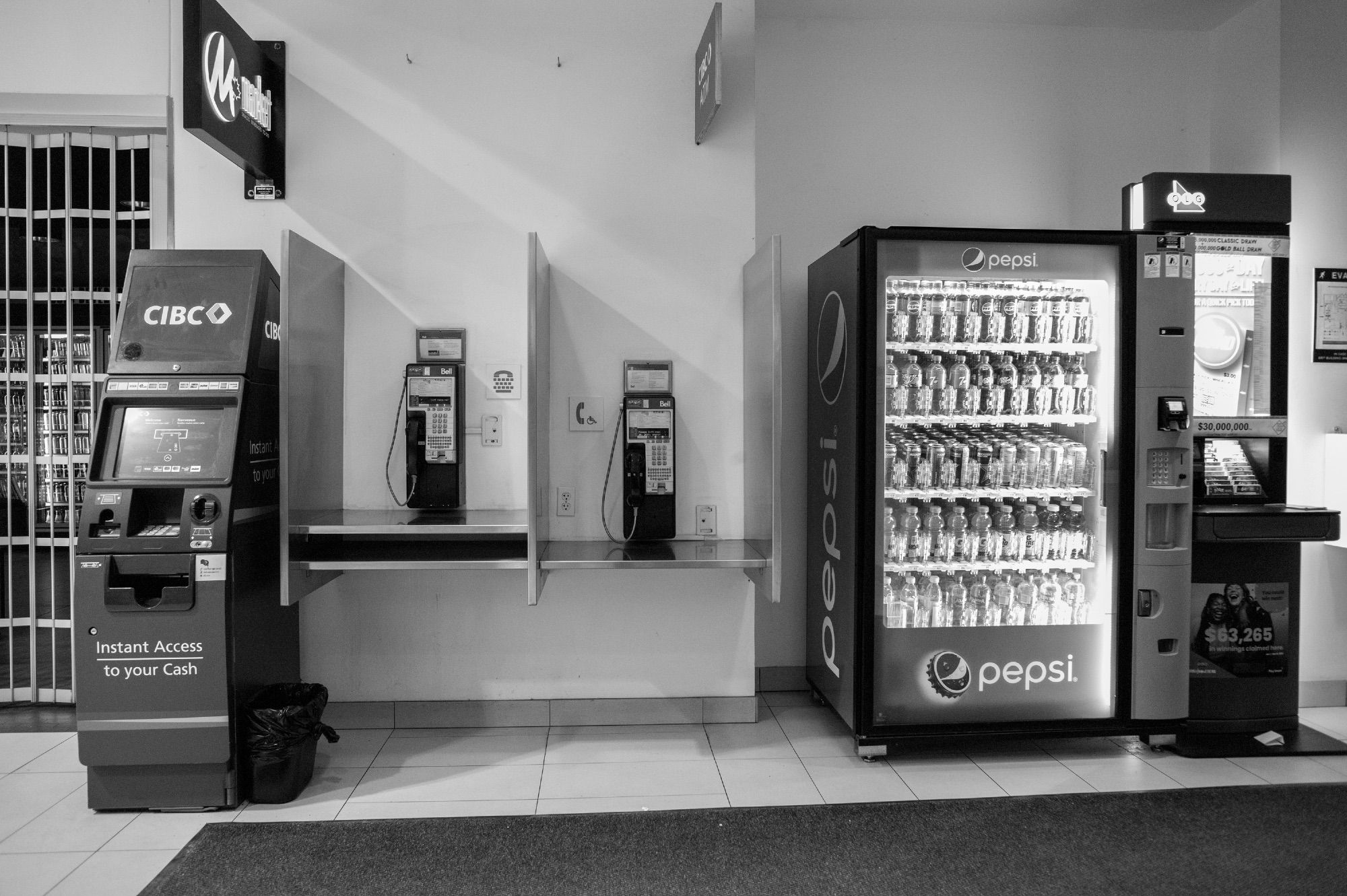 A wall showing, from left, a ATM, two pay phones one of which is lower for accessibility, a Pepsi vending machine and a terminal to check lottery tickets.