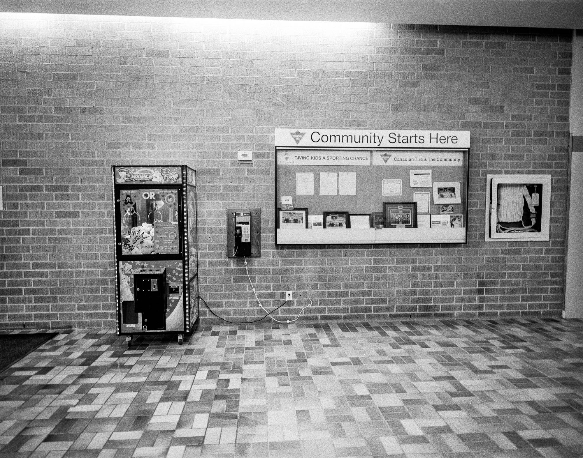 A payphone sits between a "claw" vending machine game, a glass display case with team photos and documents in it and a fire hose. A sign above the display case has a Canadian Tire logo and the message: Community Starts here. One banner in the case reads Jumpstart, giving kids a sporting chance. The other says Canadian Tire and the community. The wall is covered in 1970s style brick the floor has an intricate tile pattern of the same vintage.