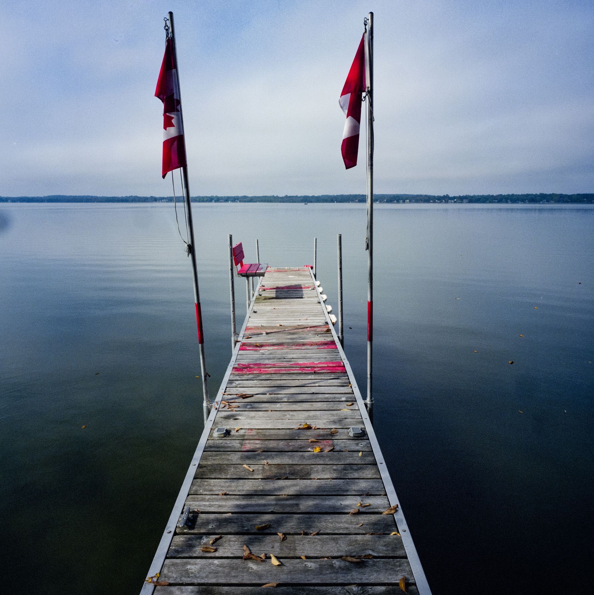 A dock on a calm morning with some fog. There are two Canadian flags flanking it. Some of the dock boards are painted red and there is a red bench built into the dock.