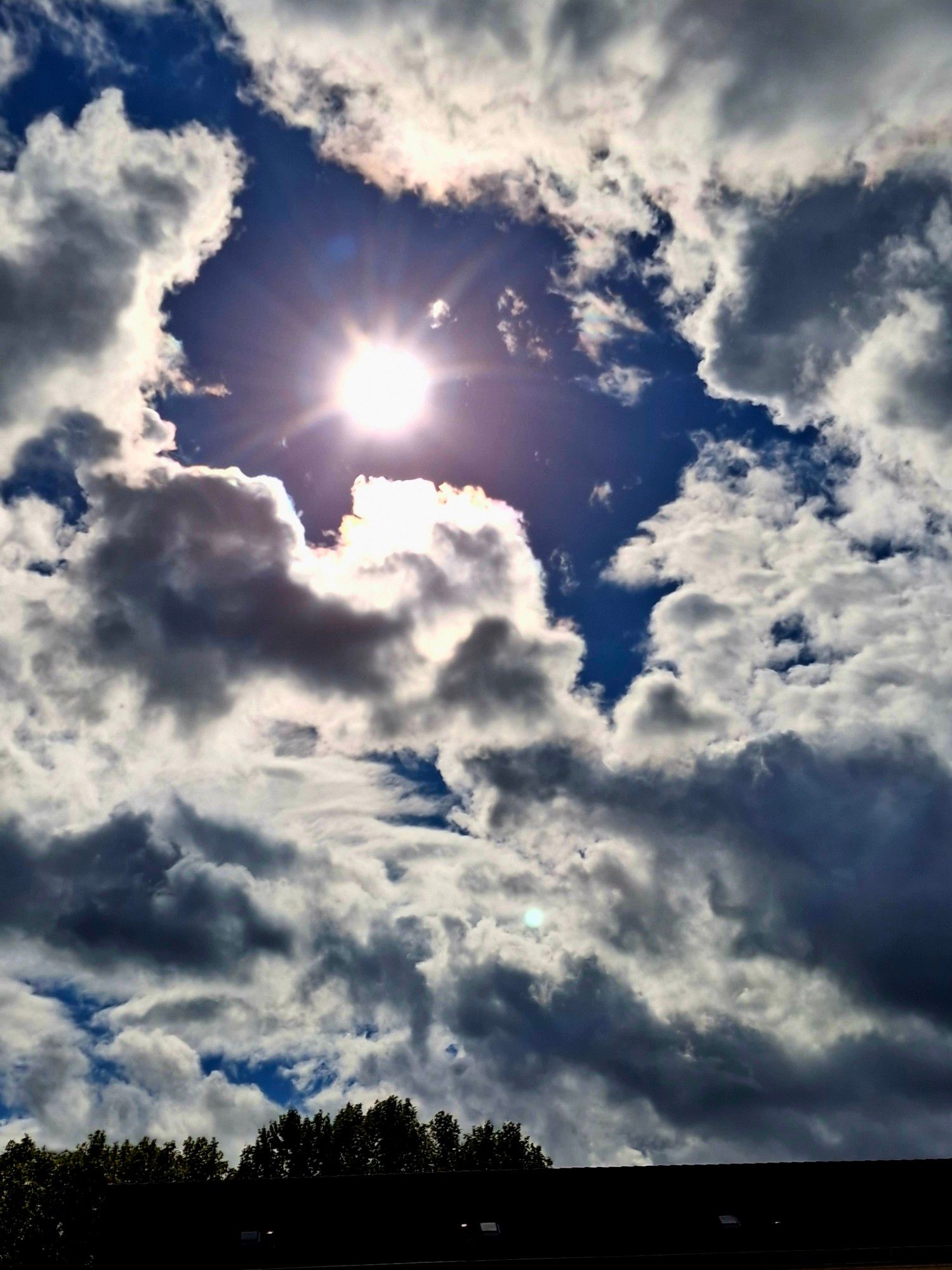 Blick aus dem Bürofenster. Baumspitzen und das Dach von einem Wohnhaus. Ganz viele Wolken und dazwischen blauer Himmel mit Sonnenschein...