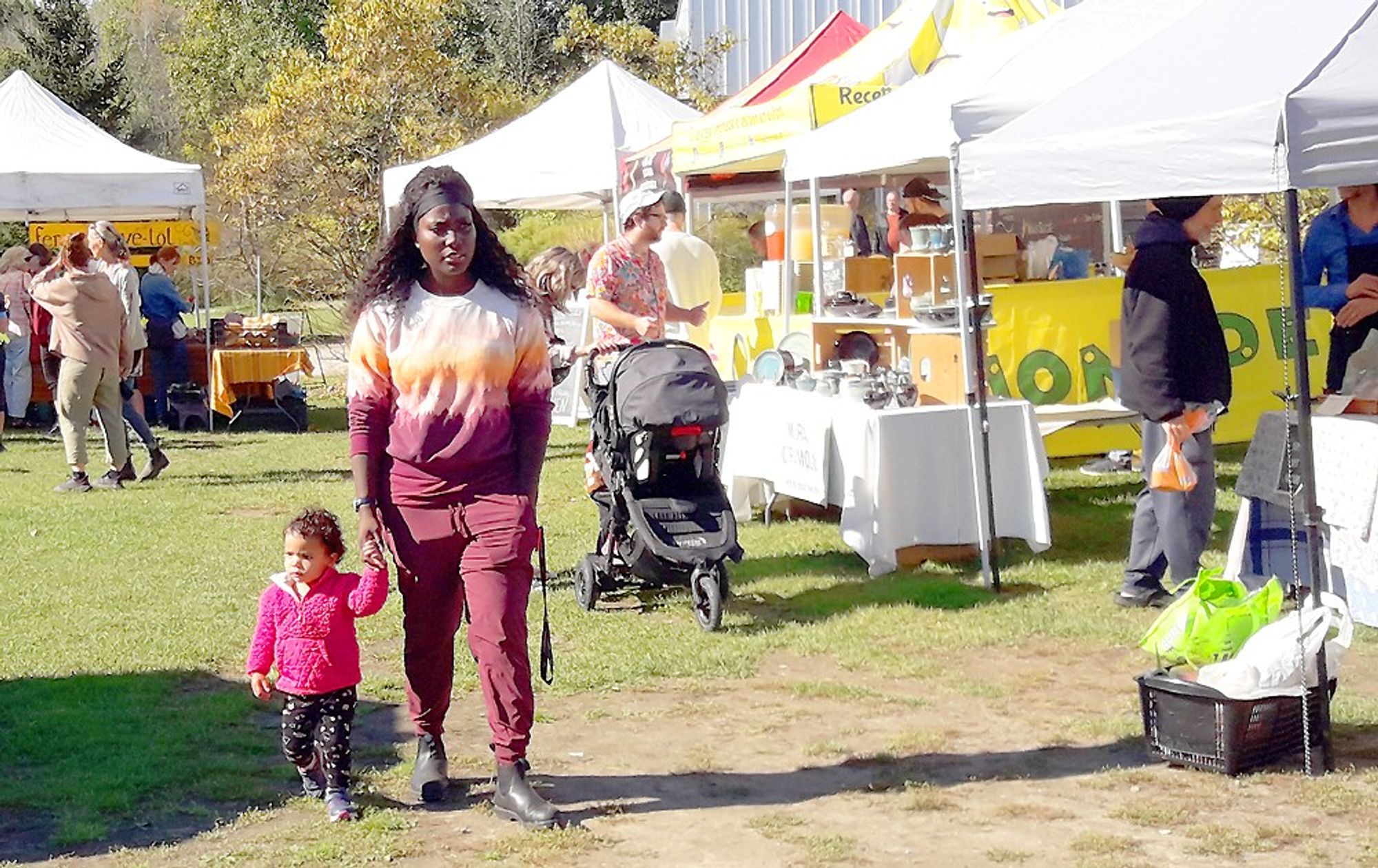 A toddler holding her mother's hand in the foreground at the left and a man pushing a baby in a carriage behind at the market in the village.