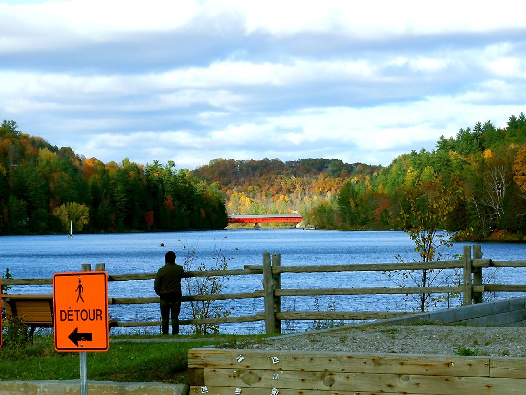 The red village covered bridge seen across the bay amidst the fall colours of the leaves in the hills behind it, while a lonely tourist is gazing at the scene standing against a split rail cedar fence.