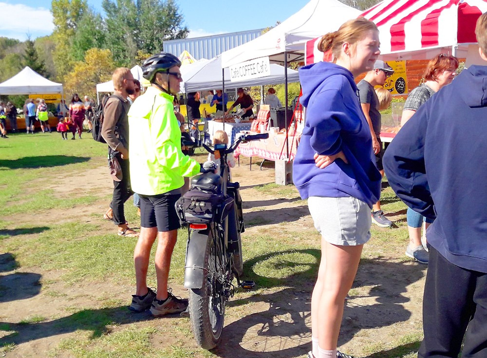 A skinny little guy in a florescent green jacket and bike helmet pushing around a gargantuan e-bike at the village market. Its size could make it pass for a motorcycle. The bike should have been parked.