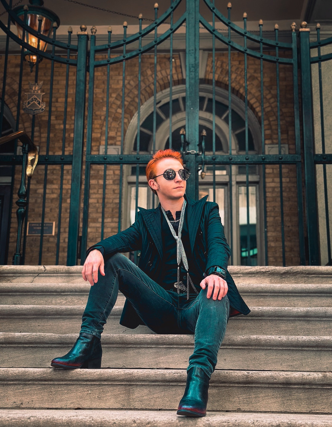 OP wearing a Crowley cosplay from Good Omens and reclining on stone steps in front of an iron gate