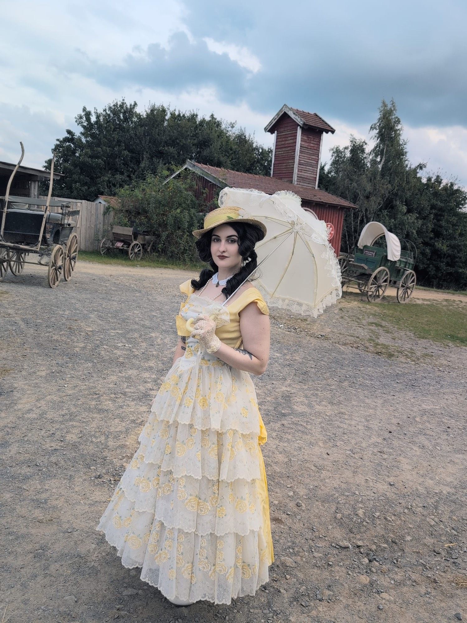 A female presenting person standing in front of a Wild West inspired scenery, and is wearing a pale yellow historical gown as well as a sun hat and a parasol.