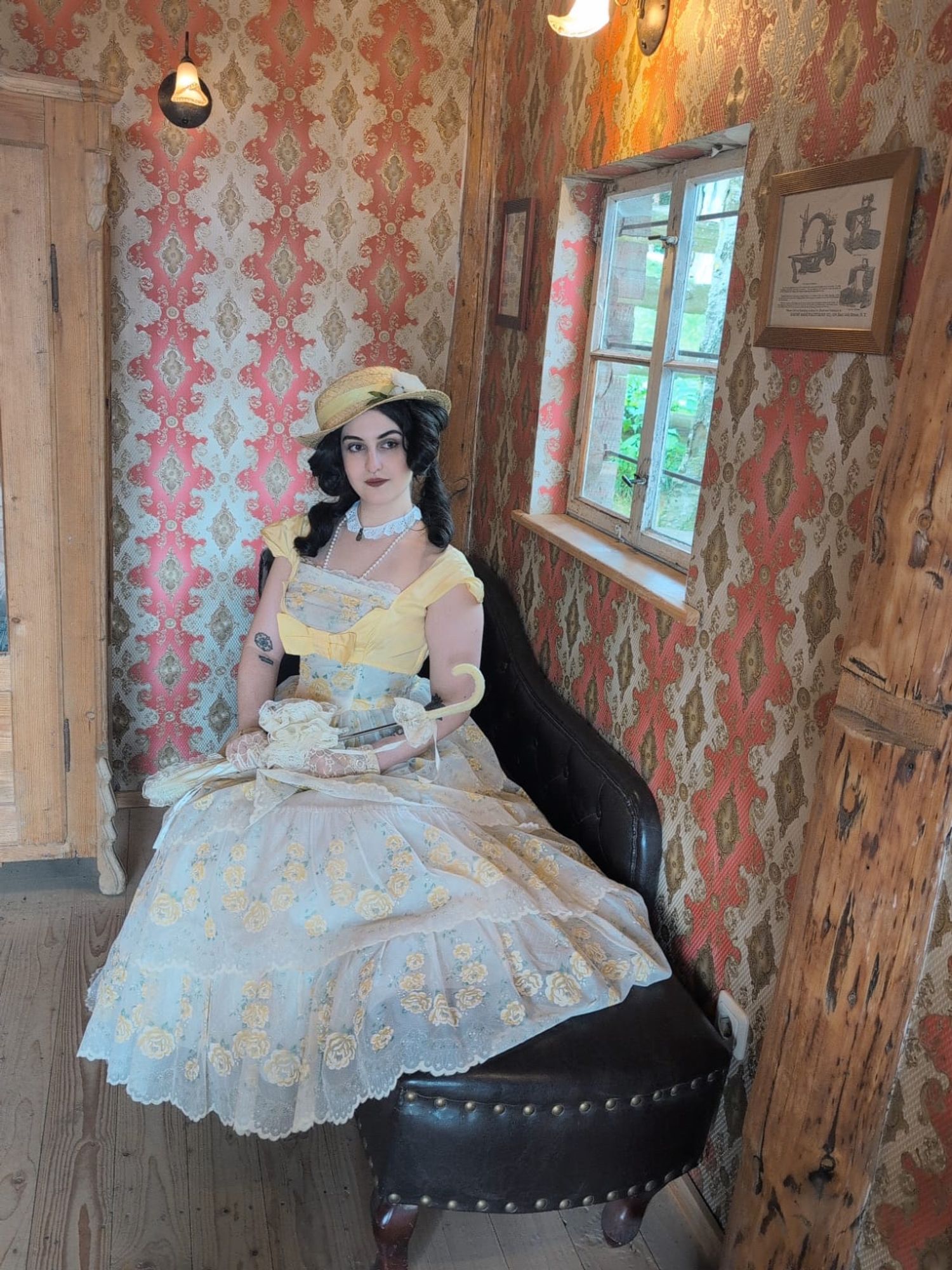 A female presenting person sitting in front of a vintage scenery, and is wearing a pale yellow historical gown as well as a sun hat and a closed parasol on their lap.