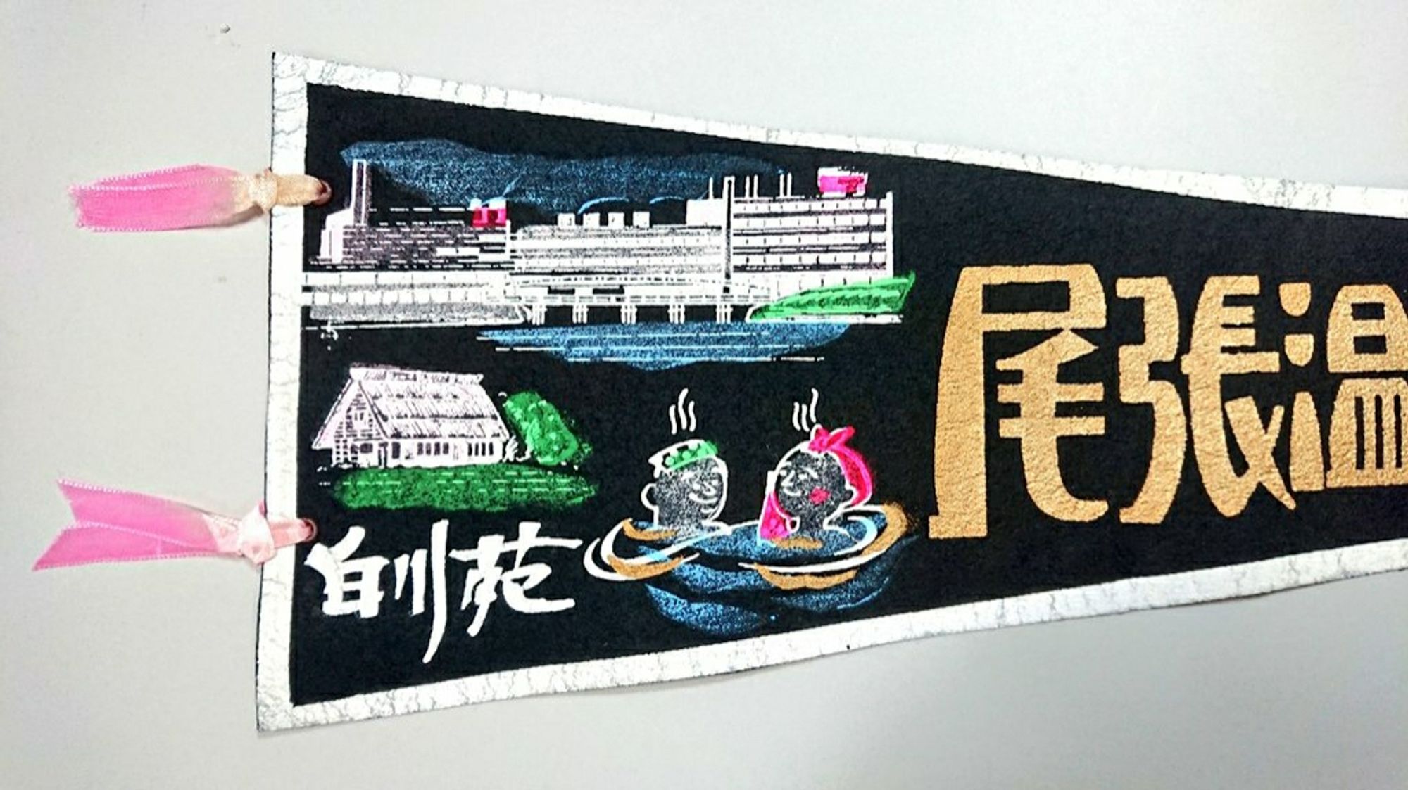 2, A pennant of Owari Onsen. It depicts a modern reinforced concrete building and two guests taking a bath comfortably with a tenugui towel on their heads.