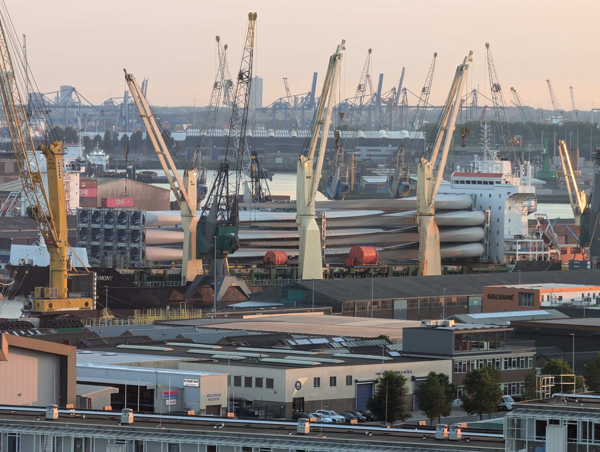 Foto van de Waalhaven met vooral veel havenkranen zichtbaar en tussen de kranen in zie je een boot liggen met op het dek ongeveer 35 wieken voor grote windmolens.