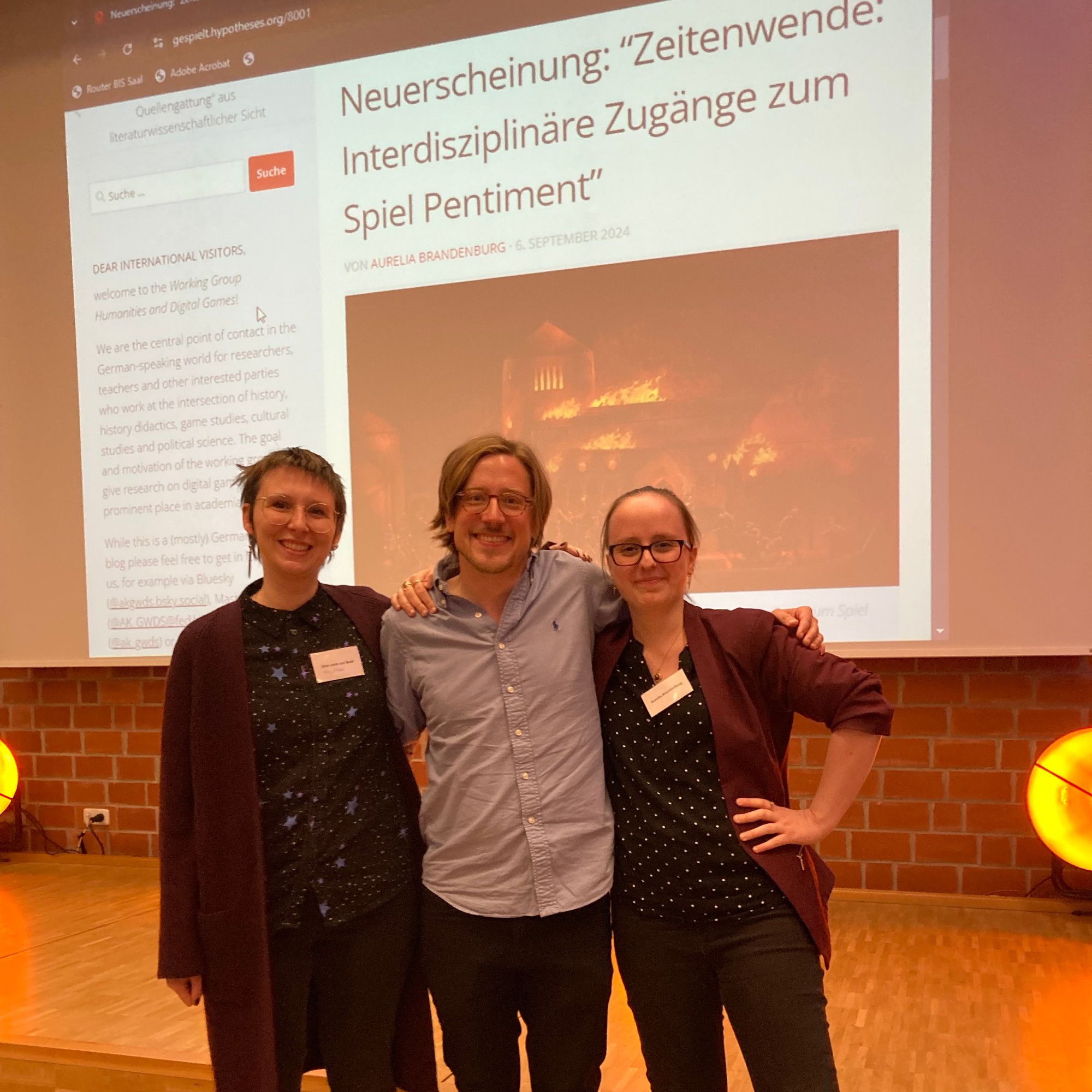 Gruppenbild mit Alan van Beek (links) & Aurelia Brandenburg auf der Tagung die "Frühe Neuzeit in Games" in Oldenburg, bei der wir den Band offiziell vorgestellt haben. Der gemeinsame Moment war unheimlich schön - nachdem wir so viel gemeinsam gewuppt haben! Im Hintergrund der AKGWDS Blog mit dem Hinweis zur Neuerscheinung und brennender Abtei.