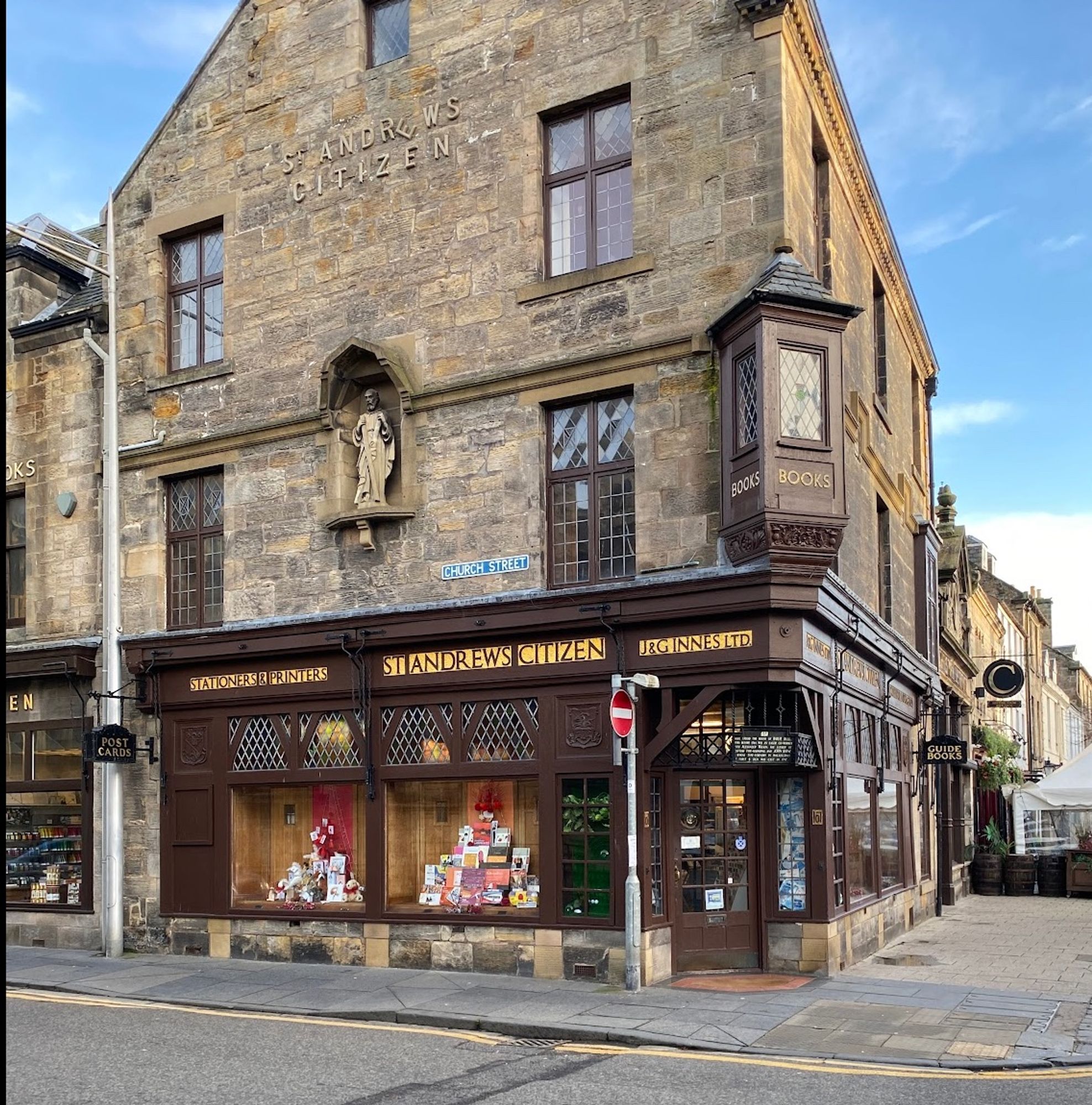 J & G Innes Ltd shop in St Andrews, Fife, on the corner of Church Street and South Street. Three-storey and attic corner building, in stone with dark brown old-fashioned shopfront. A niche at first floor level has a statue in it, the corner has a timber oriel window. All very oldee-worldee
