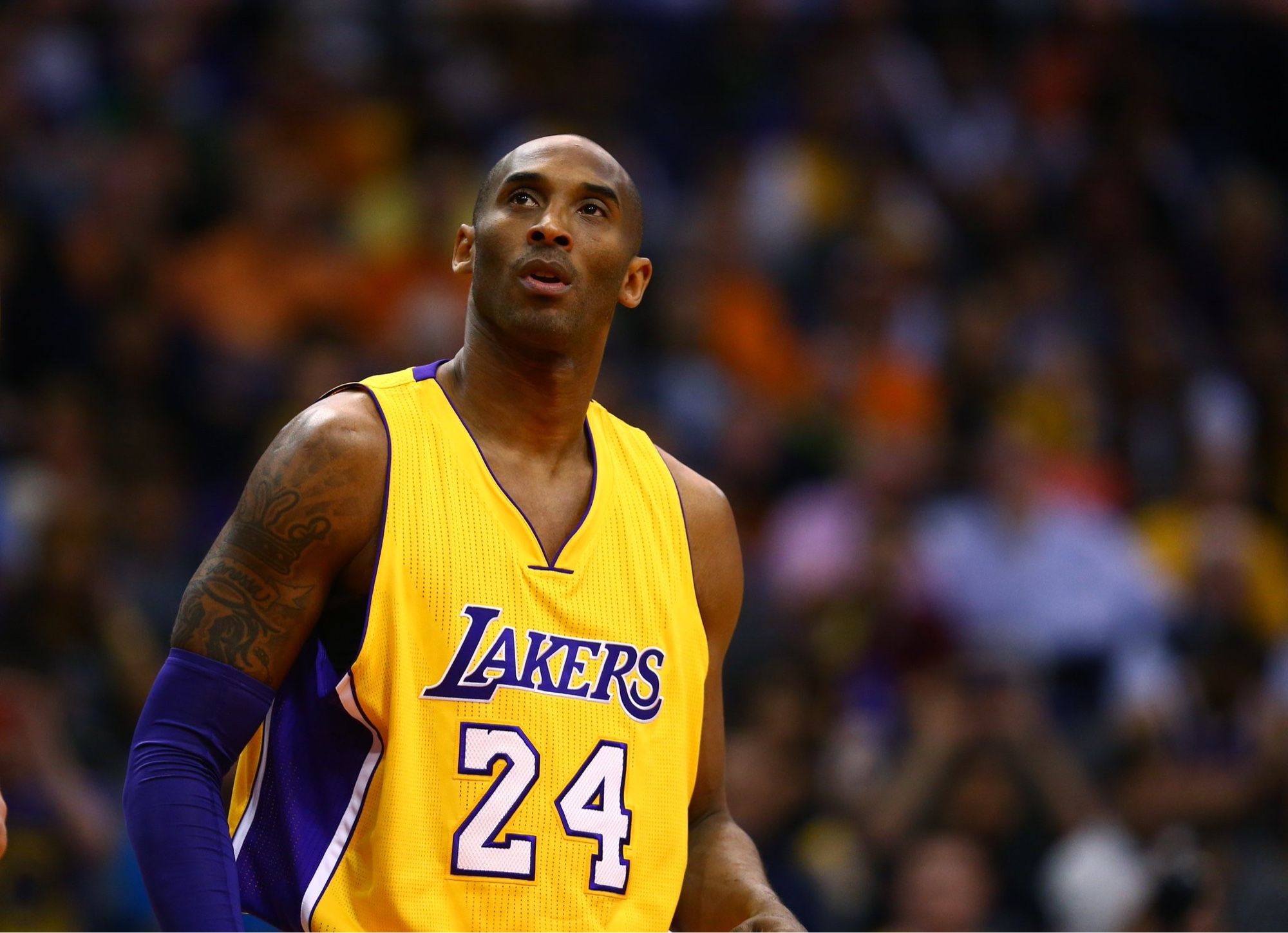 Kobe Bryant on a basketball court. He is wearing a Lakers jersey and looking up.