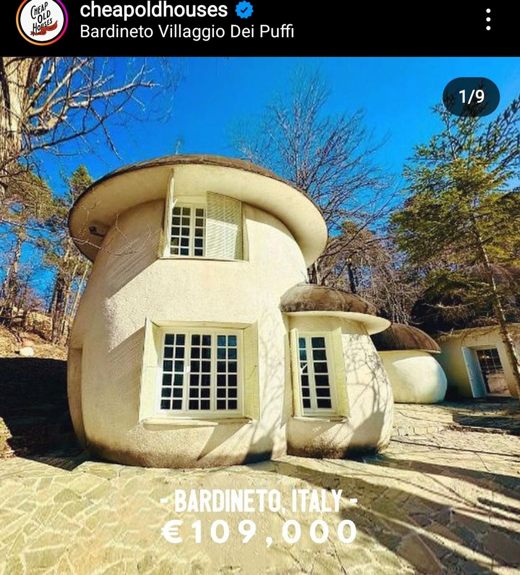 screenshot of a house for sale in Berdineto, Italy. It's in the shape of a short but thick mushroom, with a smaller mushroom built off the side, and another farther back. The walls are white, and the roof is circular brown shaped to resemble brown mushrooms. There's a few big wood paned windows, and its a pretty small home