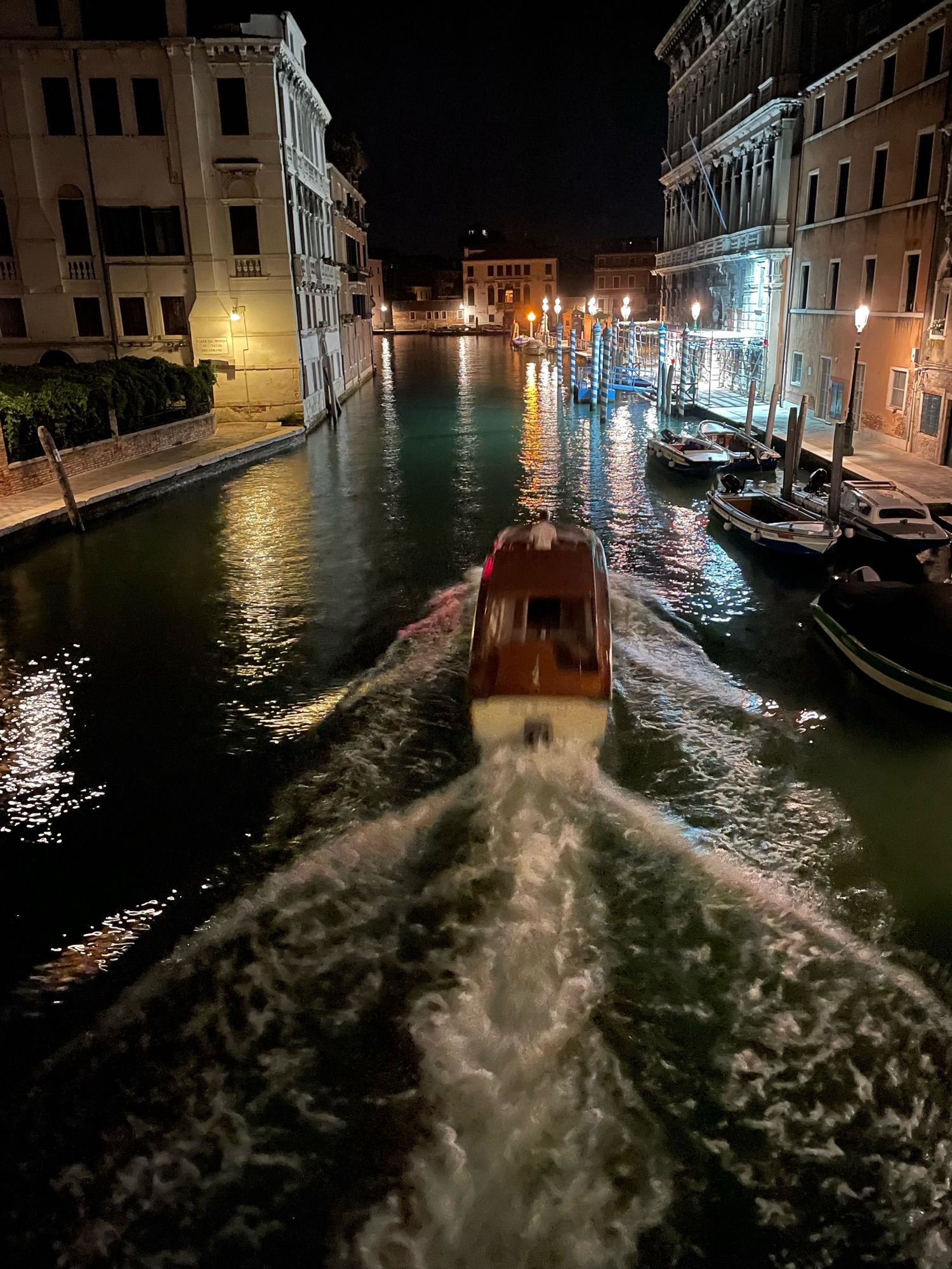 Blick auf einen Kanal in Venedig nachts, ein einzelnes Wassertaxi unterwegs auf dem Weg zum Canale Grande, der hinten im Bild zu sehen ist.