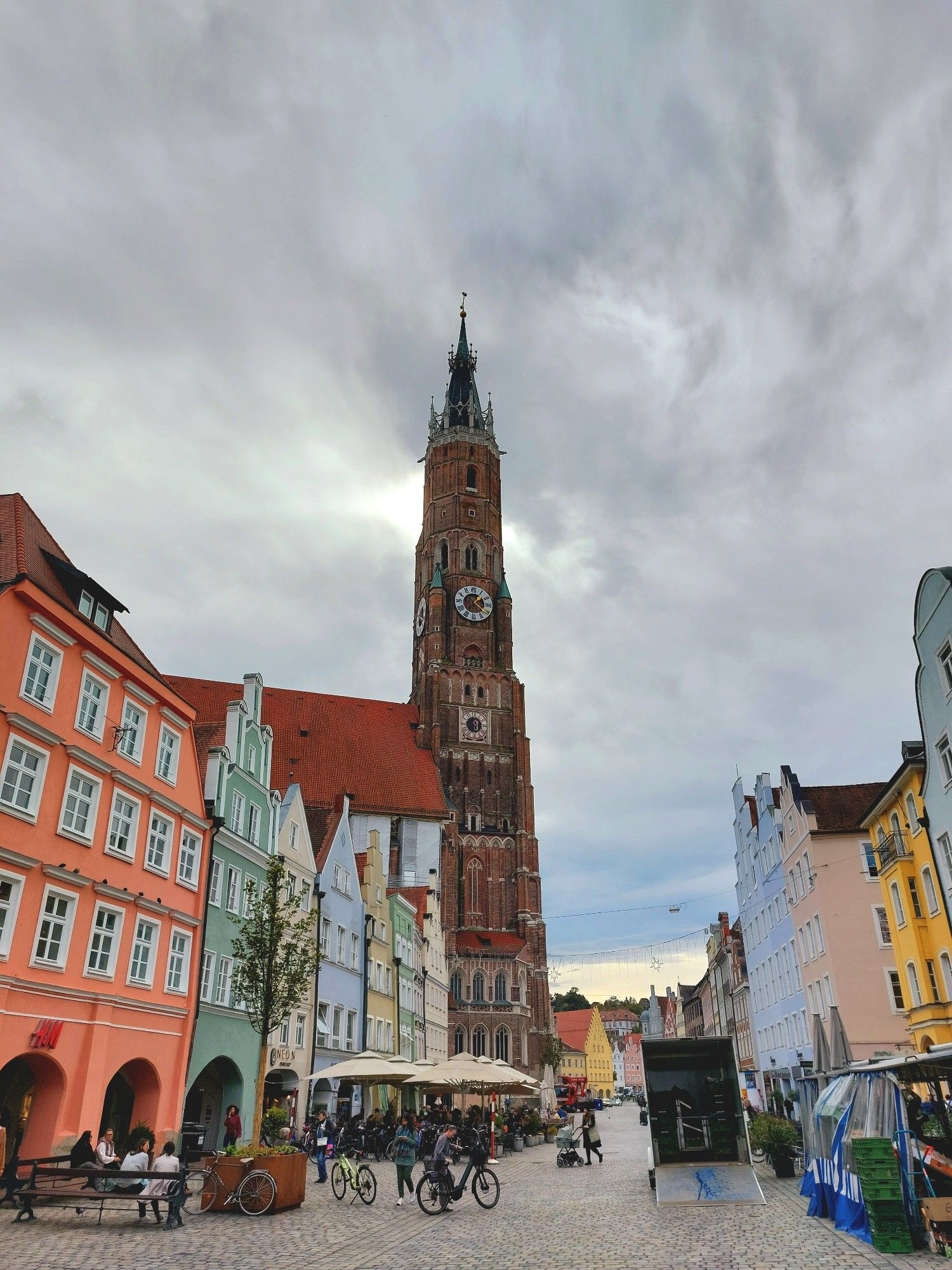 Ein großer gepflasterter Platz. Links und rechts stehen 4- und 5-geschossige alte Bürgerhäuser in den unterschiedlichsten Farben, zum Teil mit schönen Schmuckgiebeln.
In der Bildmitte erhebt sich ein aus roten Ziegeln gebauter hoher Kirchturm, der die aufgewühlten Wolken zu berühren scheint.