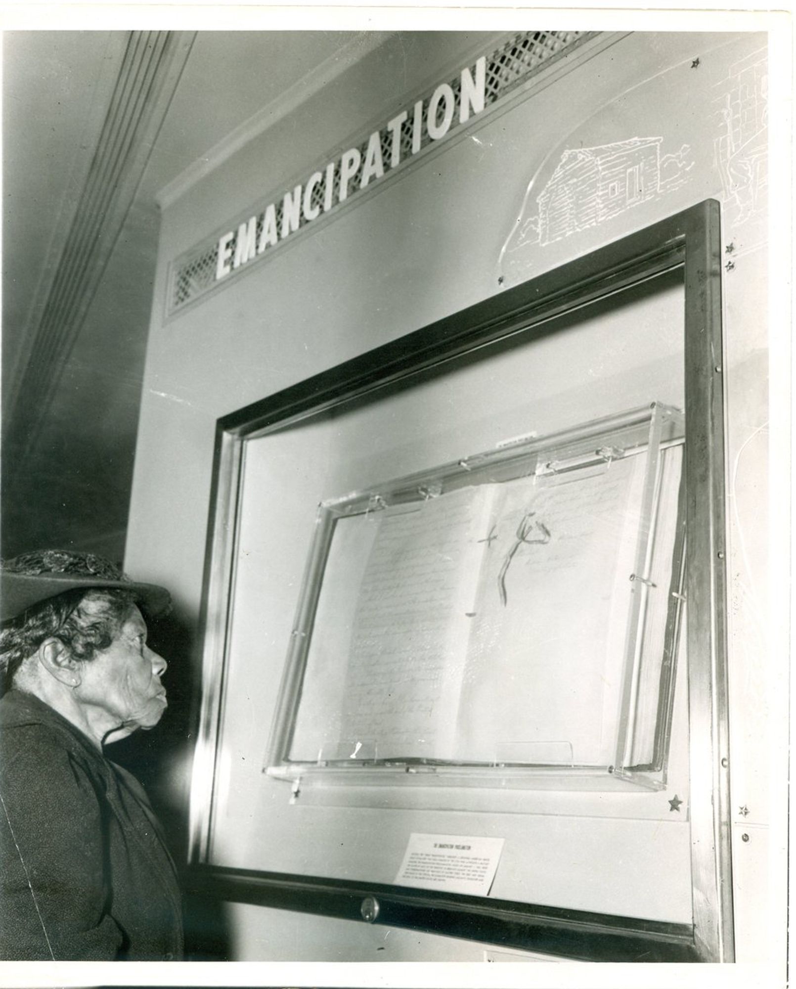 Black and white picture of an elderly Black woman wearing a hat and coat standing in from of a case with a document that contained the Emancipation Proclamation. She’s reading the text. Above the case is the word "Emancipation."