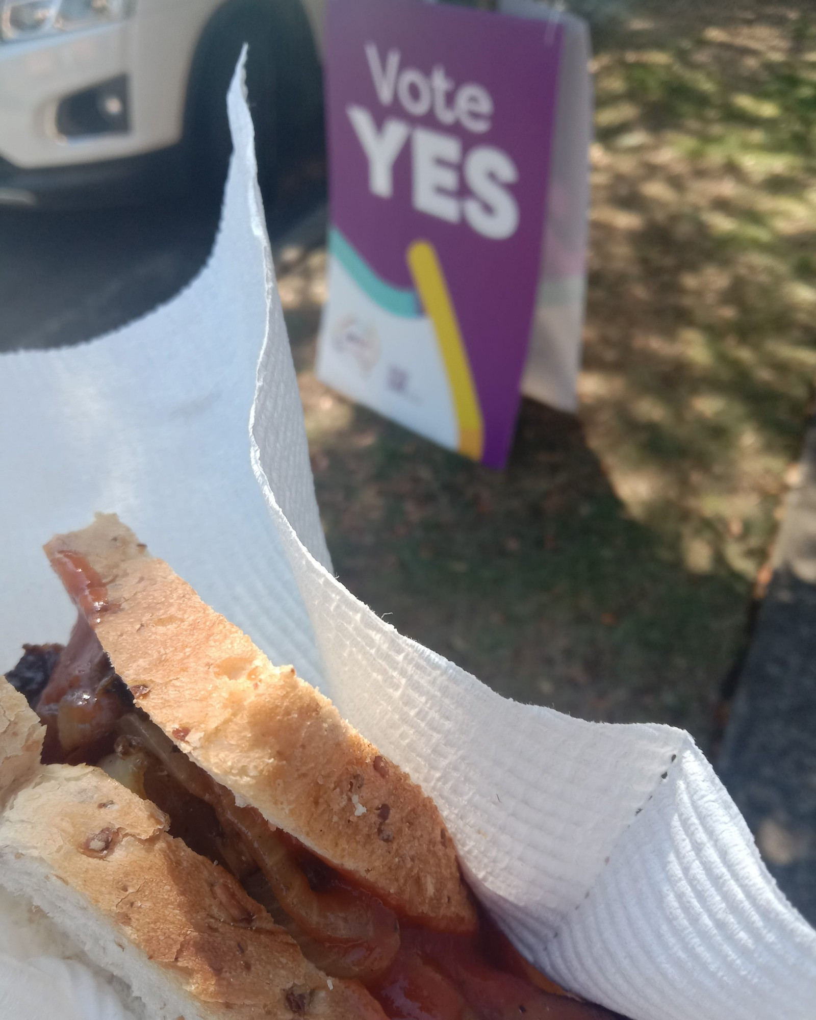 A sausage with onion and tomato sauce in a slice of multigrain bread in the foreground. A corflute with the words "Vote Yes" is in the background.