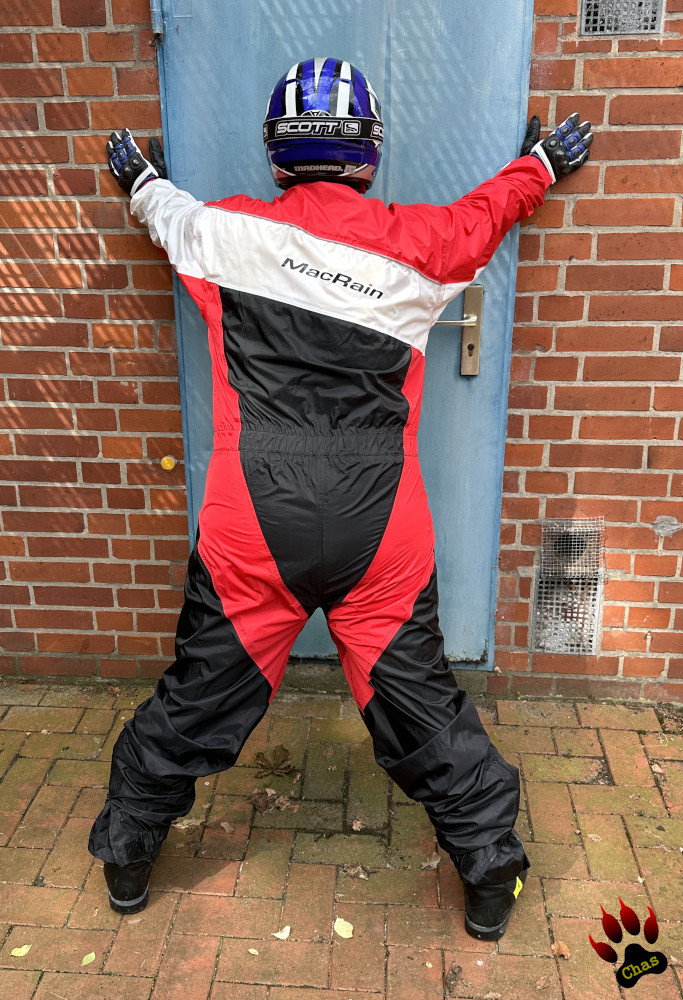 person wearing a red/black/white motorbike rainsuit, Haix boots and a motocross helmet standing at a brick wall with a blue door, leaning forward, hands on wall, feet apart