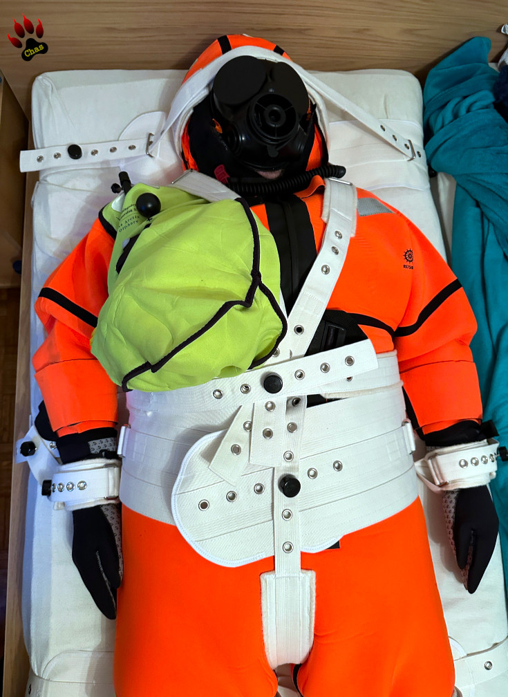 person wearing an orange neoprene immersion suit and S10 gasmask, bound to a bed using the Segufix system, upper body (torso/head) view