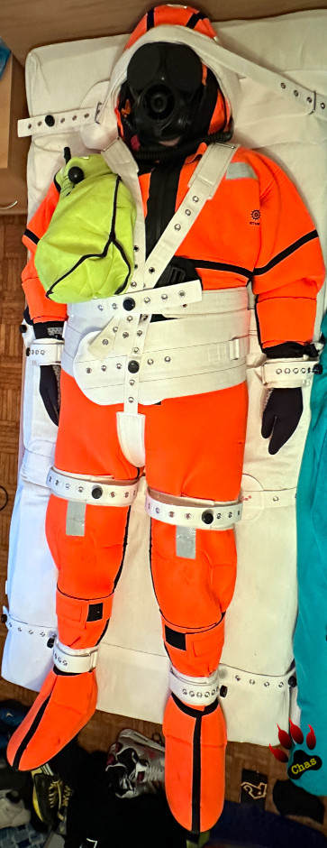 person wearing an orange neoprene immersion suit and S10 gasmask, bound to a bed using the Segufix system, full view