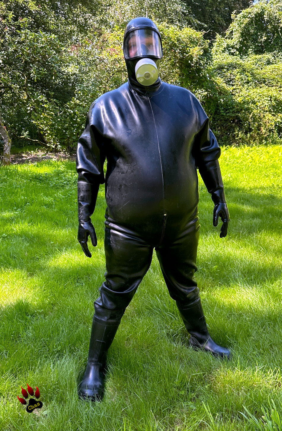 person completely covered in a heavy rubber suit with attached boots, gloves and gasmask standing in a garden, full front view