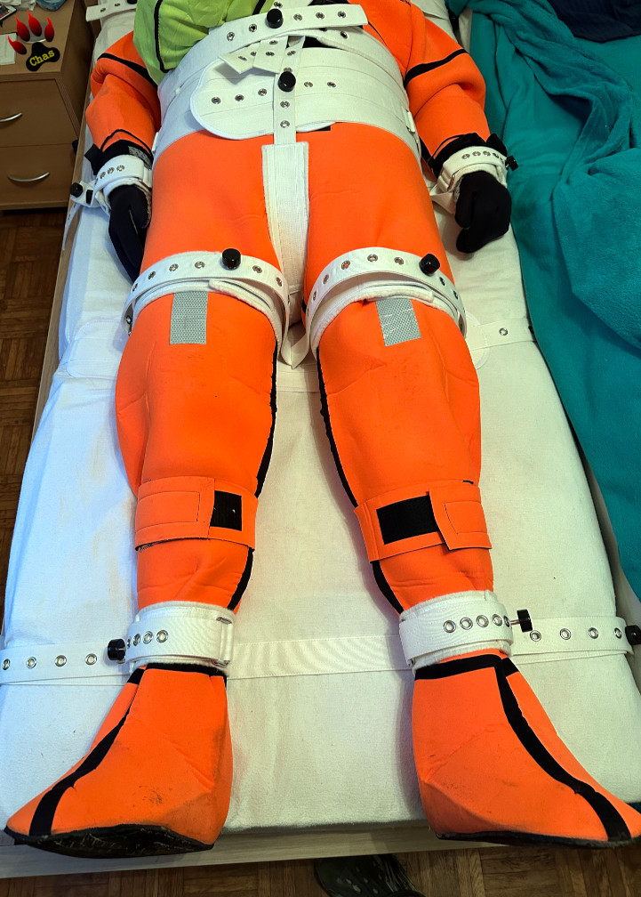 person wearing an orange neoprene immersion suit and S10 gasmask, bound to a bed using the Segufix system, lower body (torso/legs) view