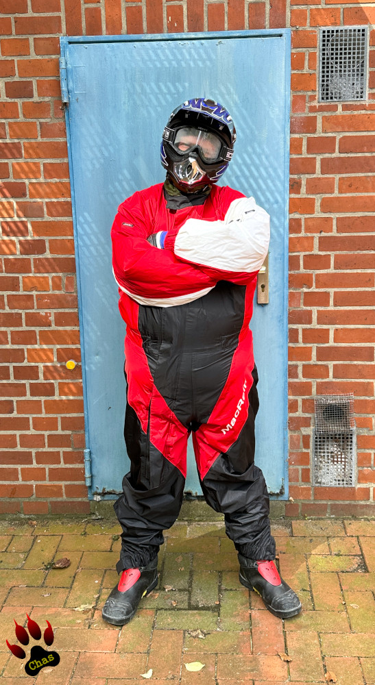 person wearing a red/black/white motorbike rainsuit, Haix boots and a motocross helmet standing outdoors in front of a brick wall with a blue door, arms crossed