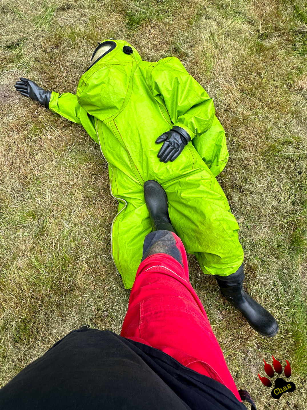 person in green hazmat suit with SCBA equipment lying on a freshly mowed lawn, another person's rubber bootet foot in his crotch