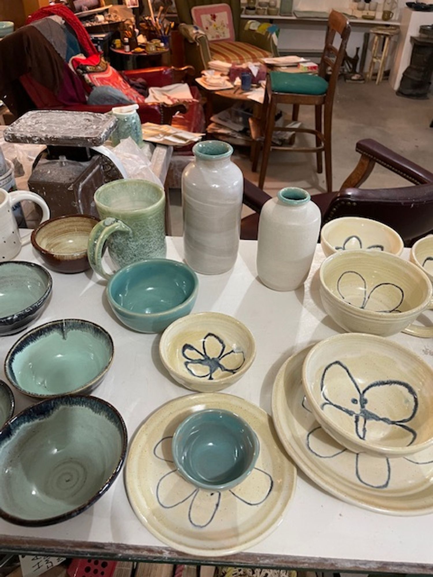 An array of wheel-thrown ceramics, including plates (ivory with a blue pattern), bowls (turquoise and black), vases (white and turquoise), plus a green speckled mug and a honey-colored bowl.