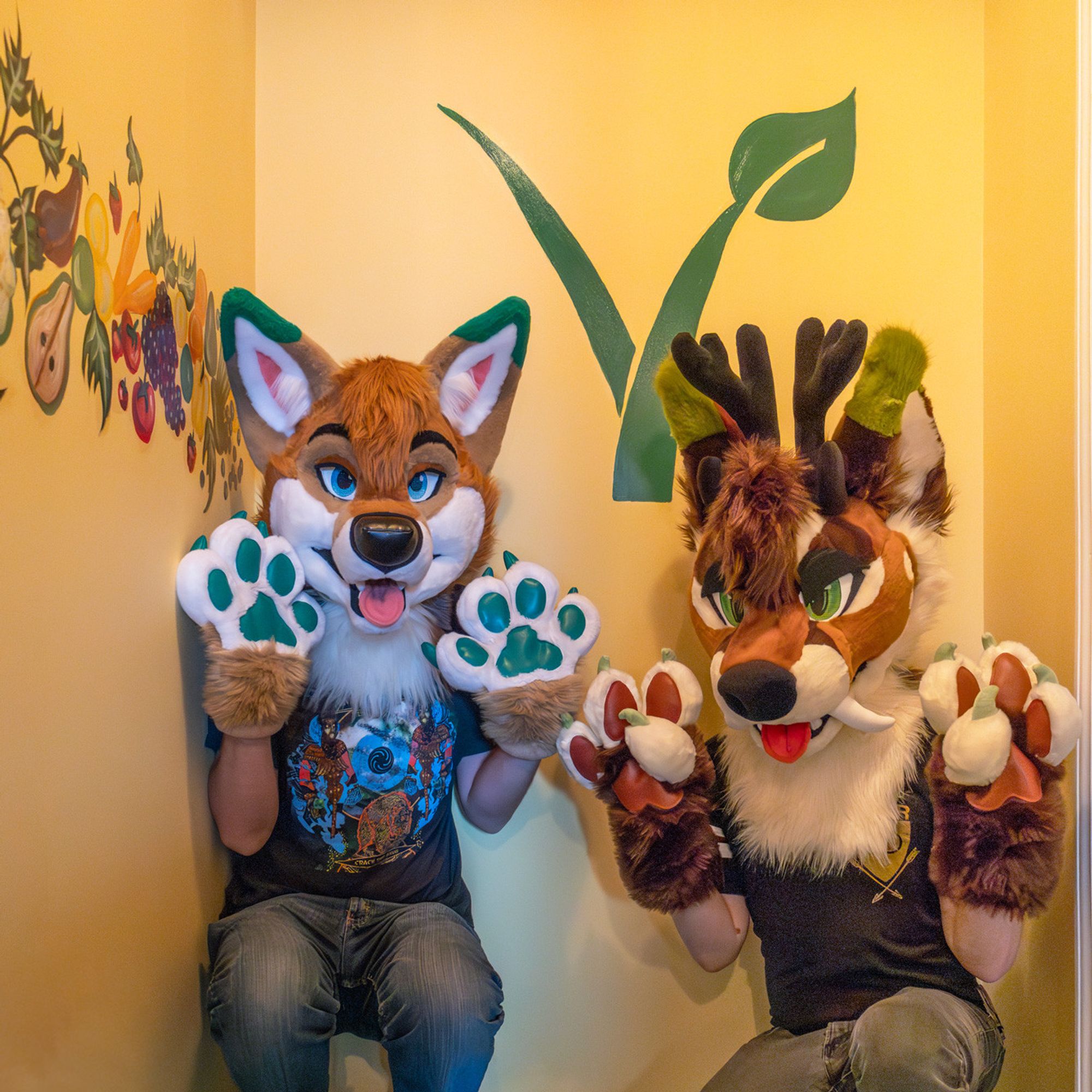 A coyote and fox-deer fursuiter crouched in front of a mural with fruit and a large green vegan V symbol holding their paws up showing their beans.