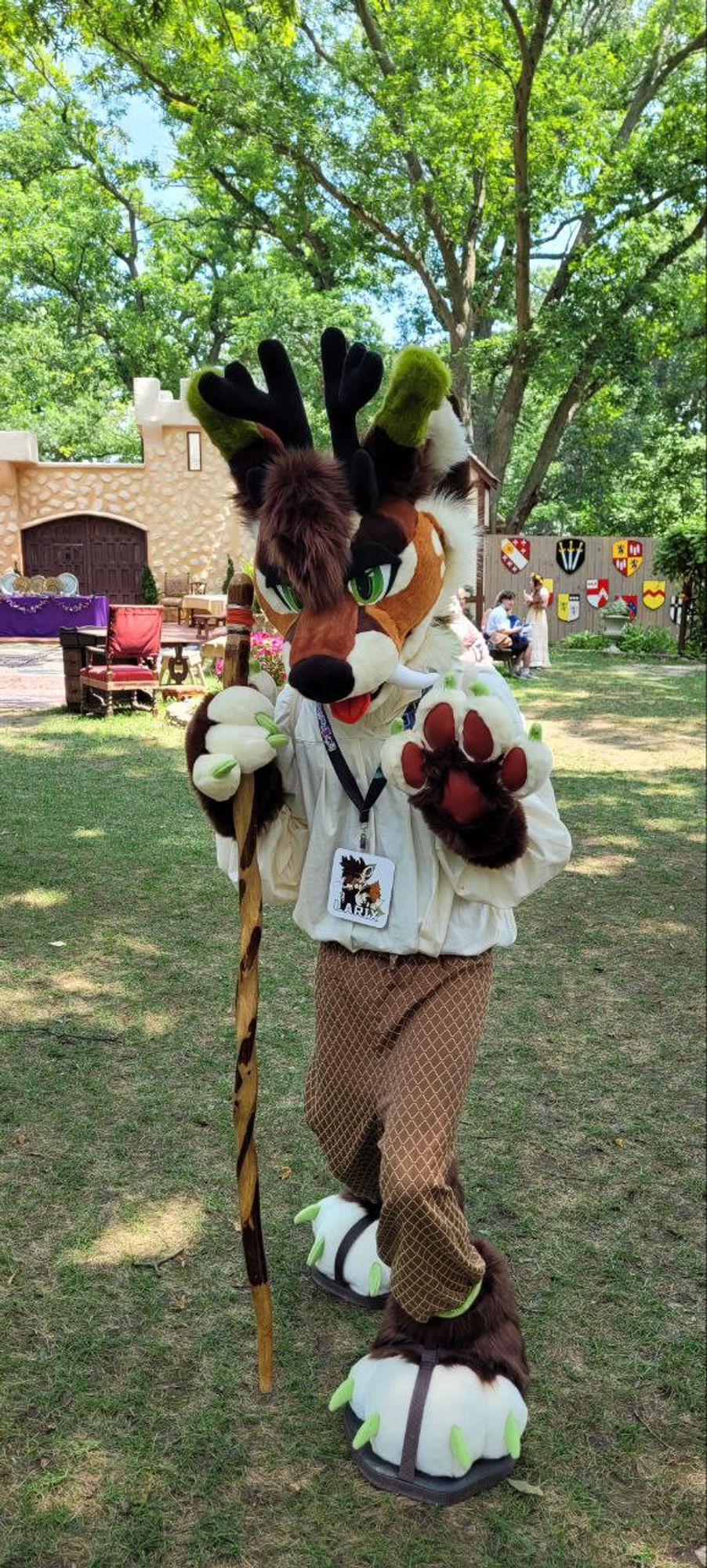 An orange, brown and green fox-deer fursuiter  holding a wooden walking stick posing in a grassy field at a renaissance faire.