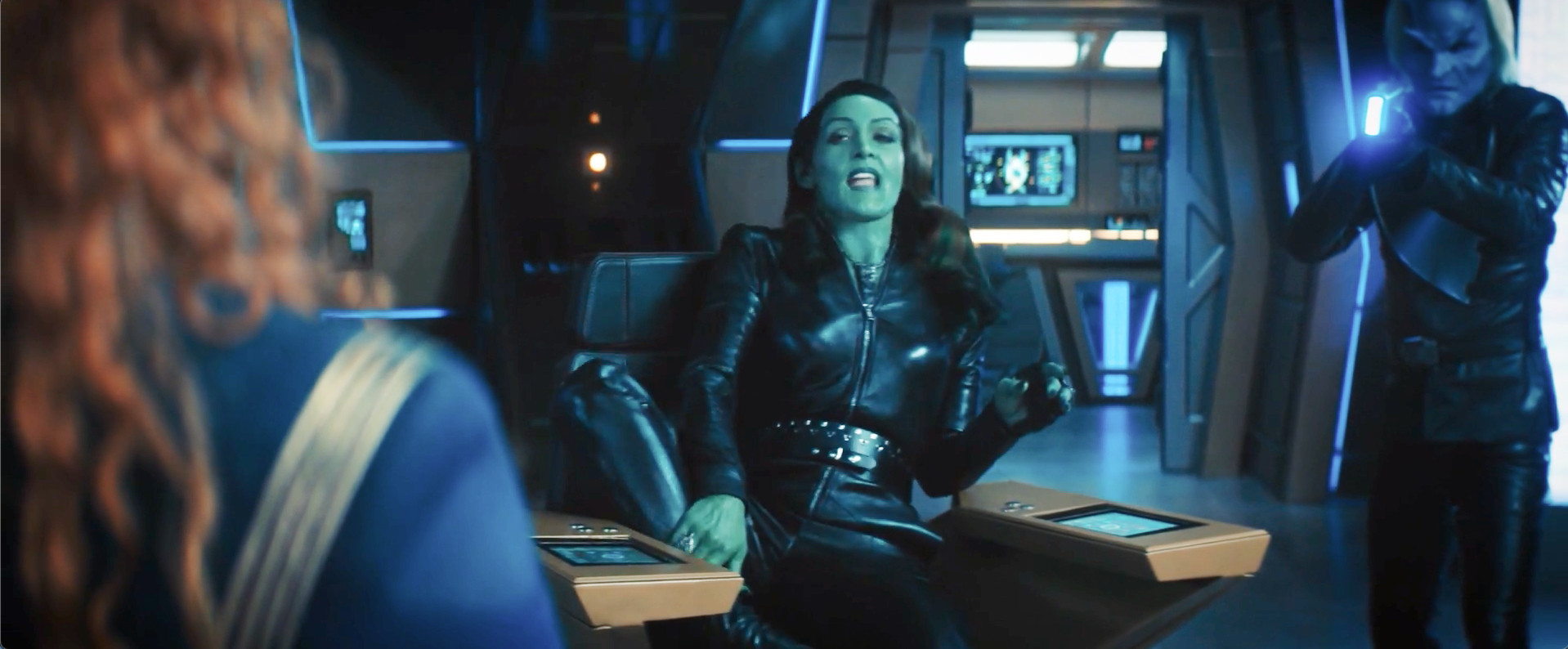 A green-skinned Orion woman is in the Captain's chair of a starship. She has contorted her legs in a traditionally "bisexual" pose that will almost certainly result in a nasty cramp.