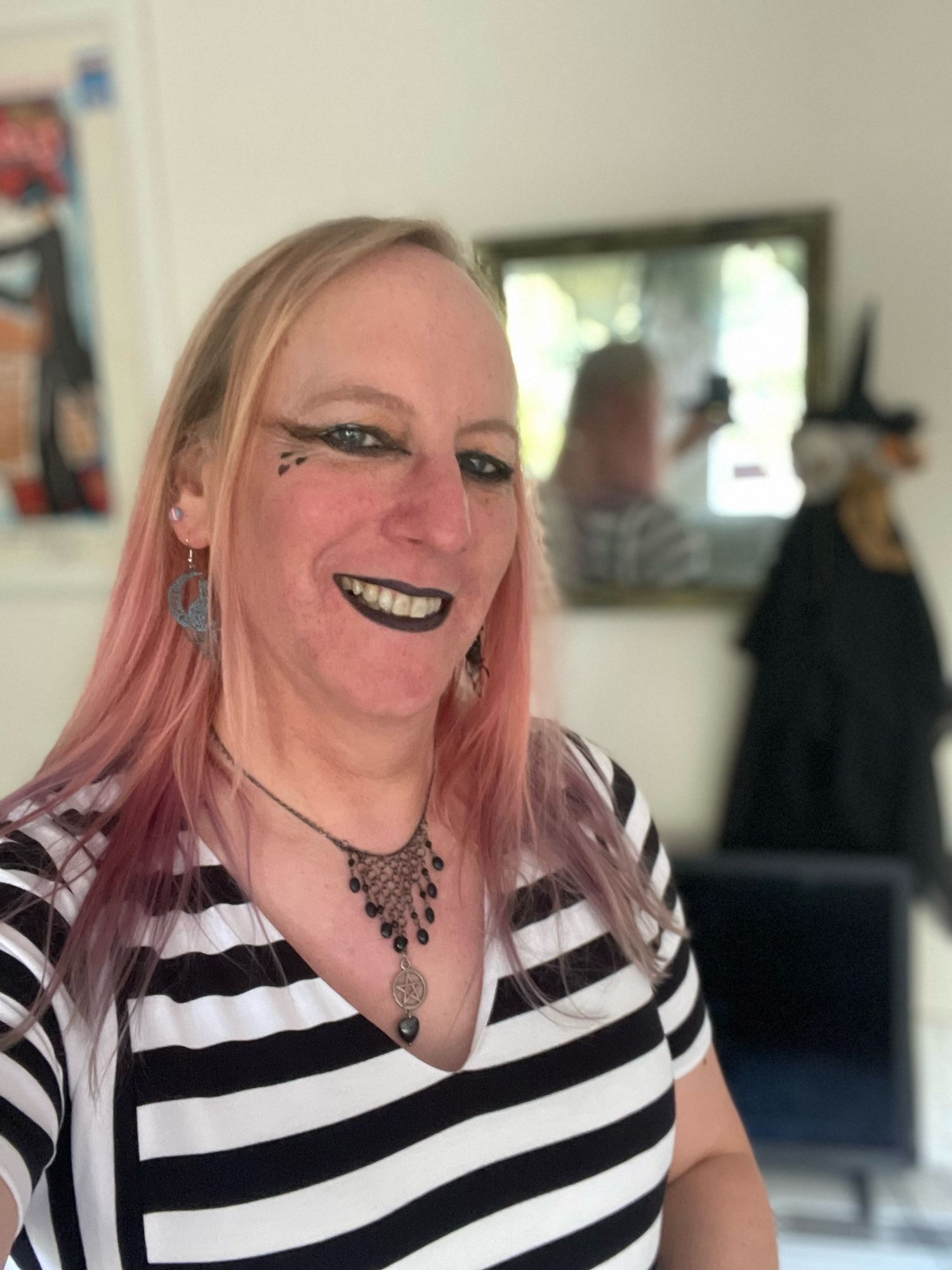 A selfie of me standing in front of a small mirror with a three-headed witch doll in front of it. I’m wearing a black and white striped dress and have black lipstick.