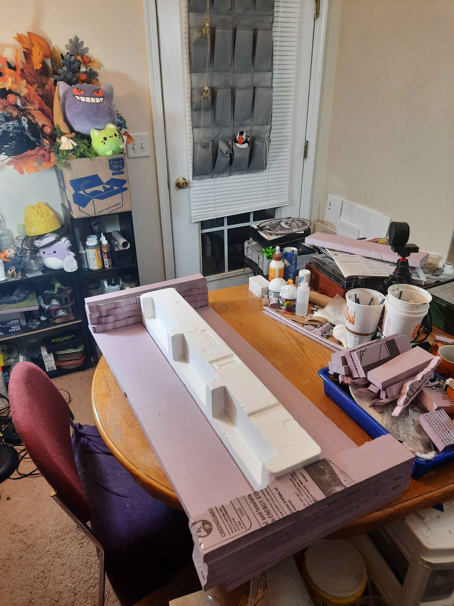 A very messy work table cluttered with a variety of different paint brushes, foam scrap, and over loaded shelves stuffed with crafting supplies. And a bridge of styrofoam on a board of insulation foam flanked by two river banks, also made of insulation foam. The banks are stacked four layers deep to match the bridge. The bridge itself is made from packing styrofoam from a tv I got who knows how long ago.