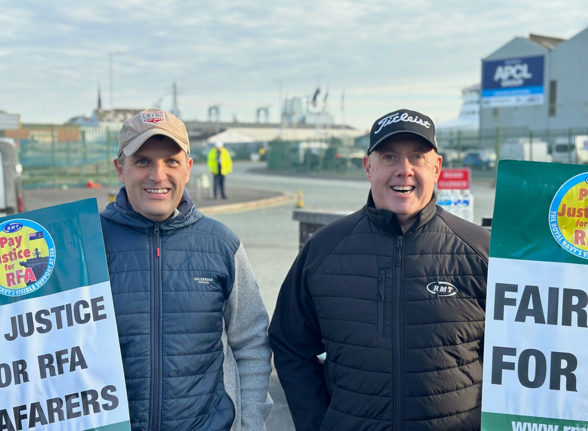 Two RMT members wearing baseball caps smile for the camera outside the Cammell Laird shipyards in Birkenhead. Both our holding placards demanding fair pay.