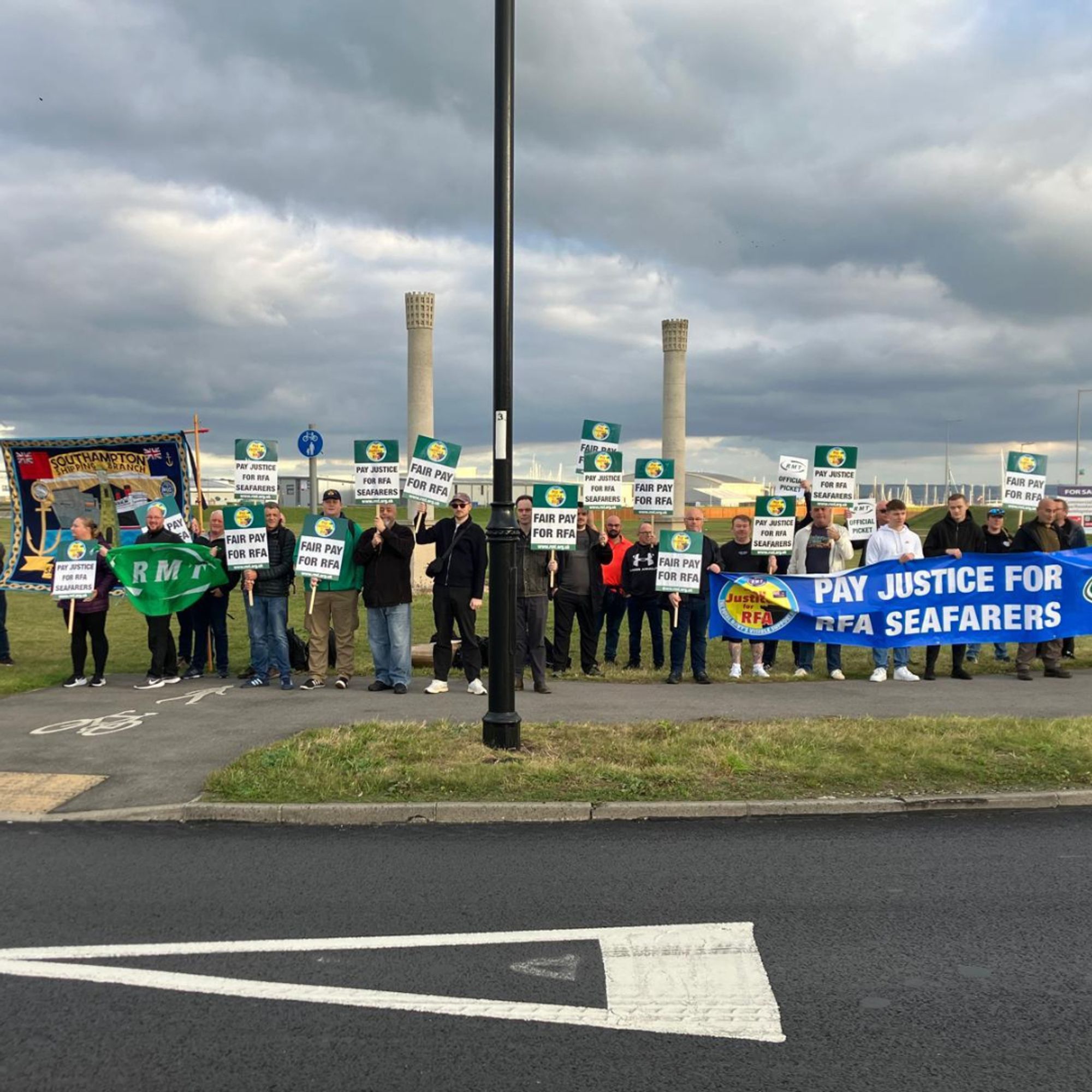 RMT Royal Fleet Auxiliary Seafarers standing outside the docks at Portland under grey, cloudy skies holding placards and banners that read 'Pay Justice For RFA Seafarers'.