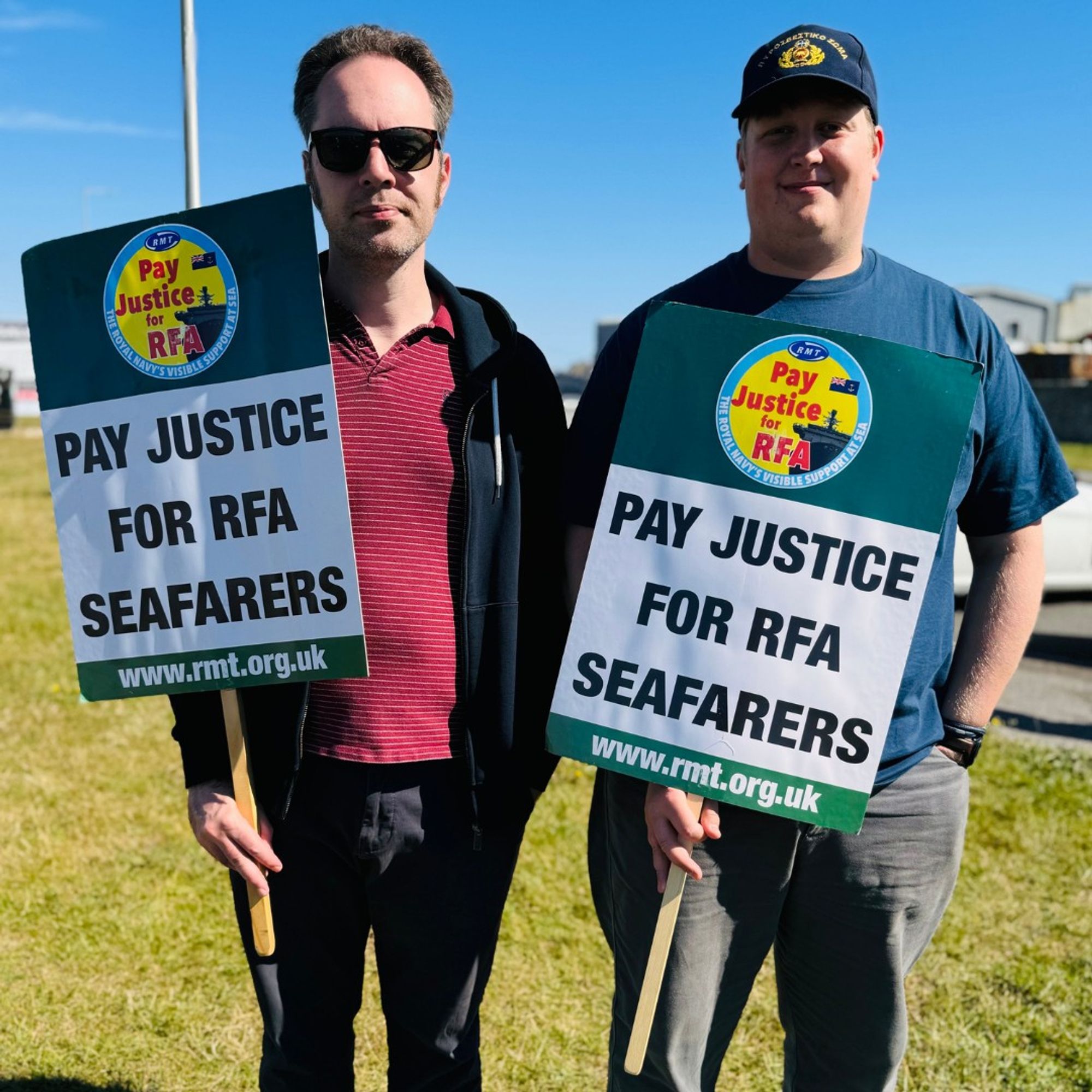 Two RMT RFA Seafarer members holding placards that 'Pay Justice For RFA Seafarers' stand on grass field under bright, blue skies.