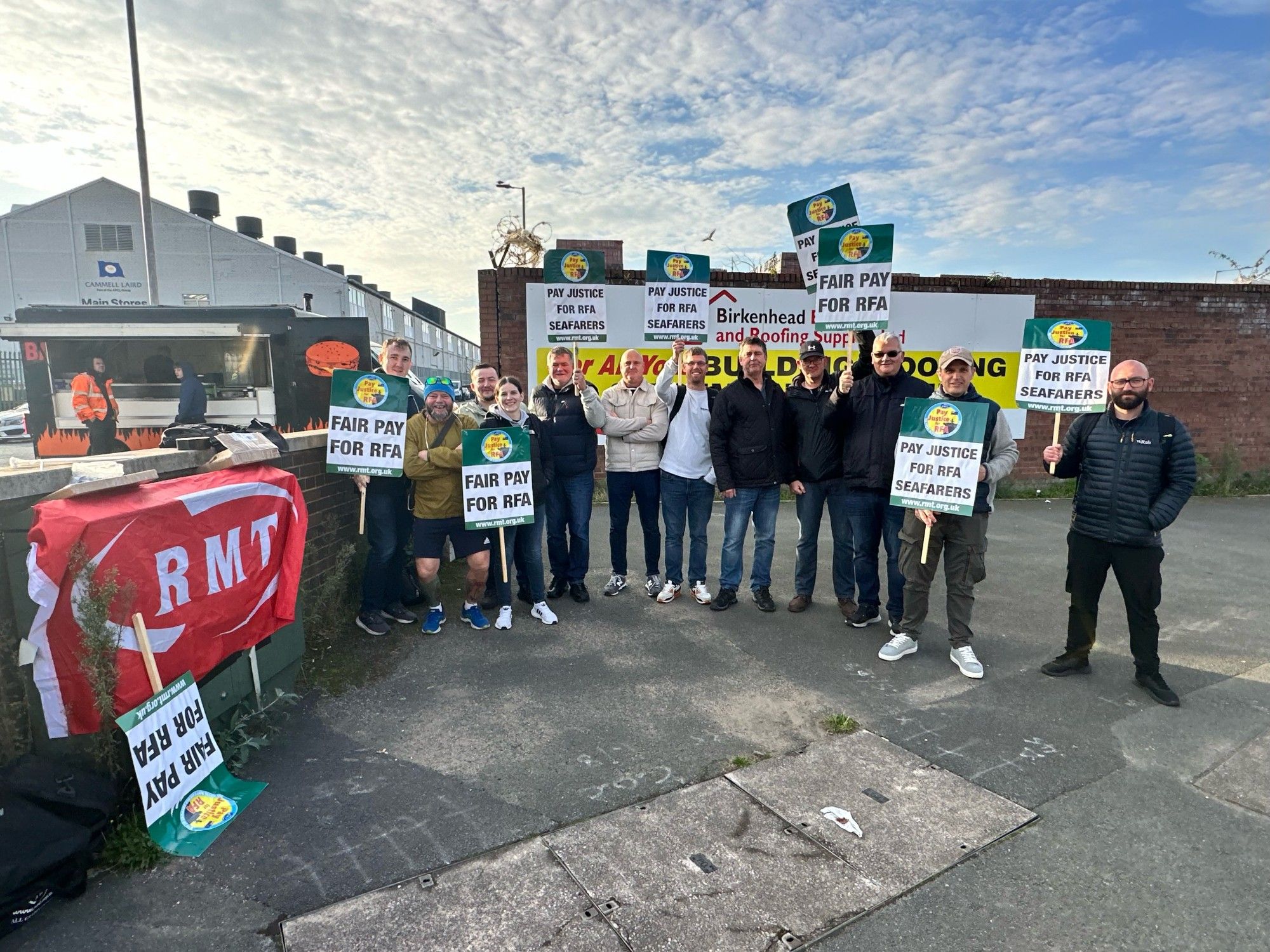 RMT members on the picket line at Cammell Laird shipyards in Birkenhead stand under blue skies holding placards that read 'Pay Justice For RFA Seafarers'.