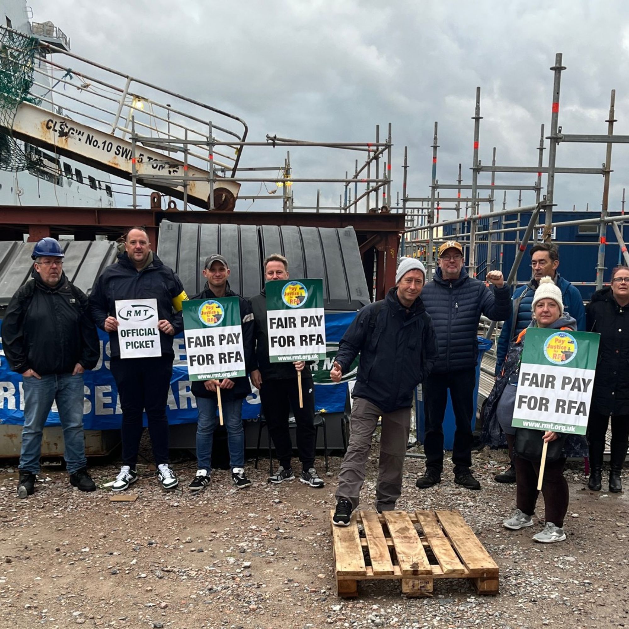 RMT members stand under leaden skies at the Cammel Laird shipyard in Birkenhead. Behind them is the gangplank to the RFA vessel Tideforce whilst our members are pictured holding placards that read 'Fair Pay For RFA' with one stands with one foot on a pallet.