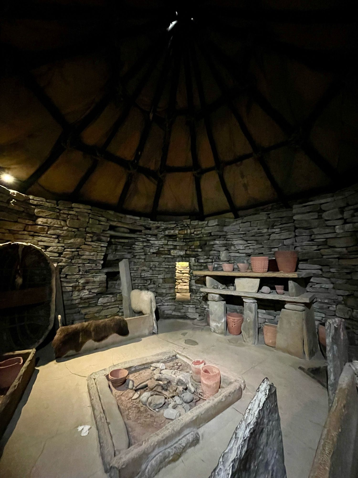 Inside a stone walled house with stone furniture and ceramic pots. A three-pillared stone shelving unit stands against the far wall, and a square stone-framed fireplace lies in the centre of the floor