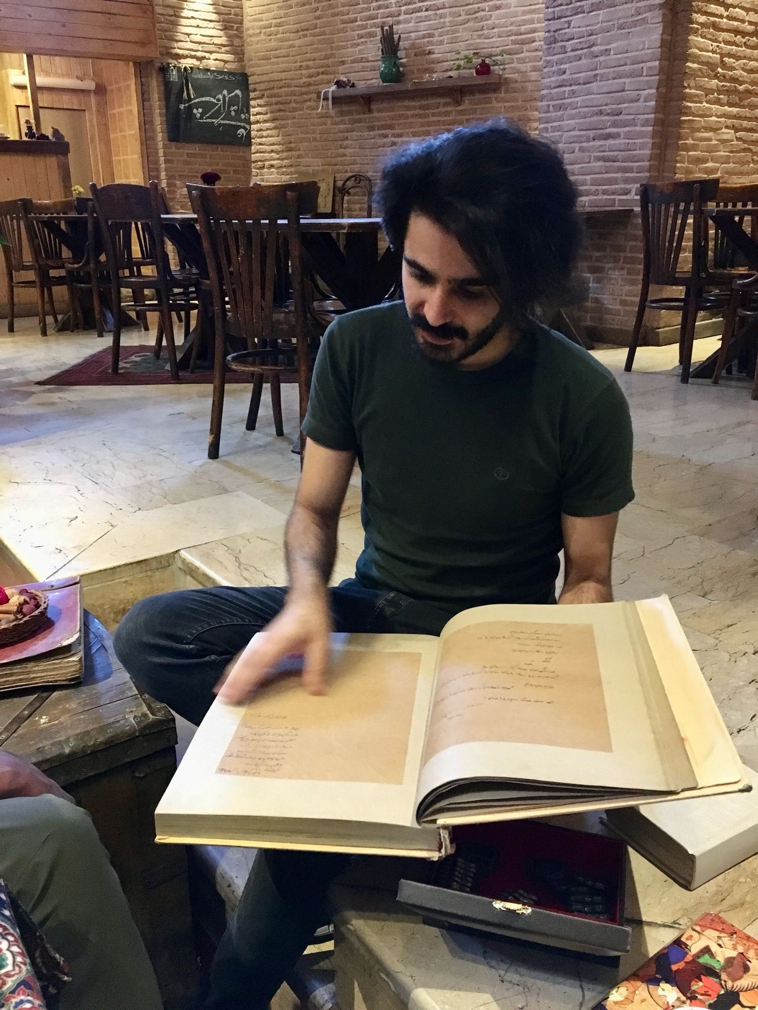 Man with curly black hair turning the pages of a large book; marble floor; brown wooden chairs and tables