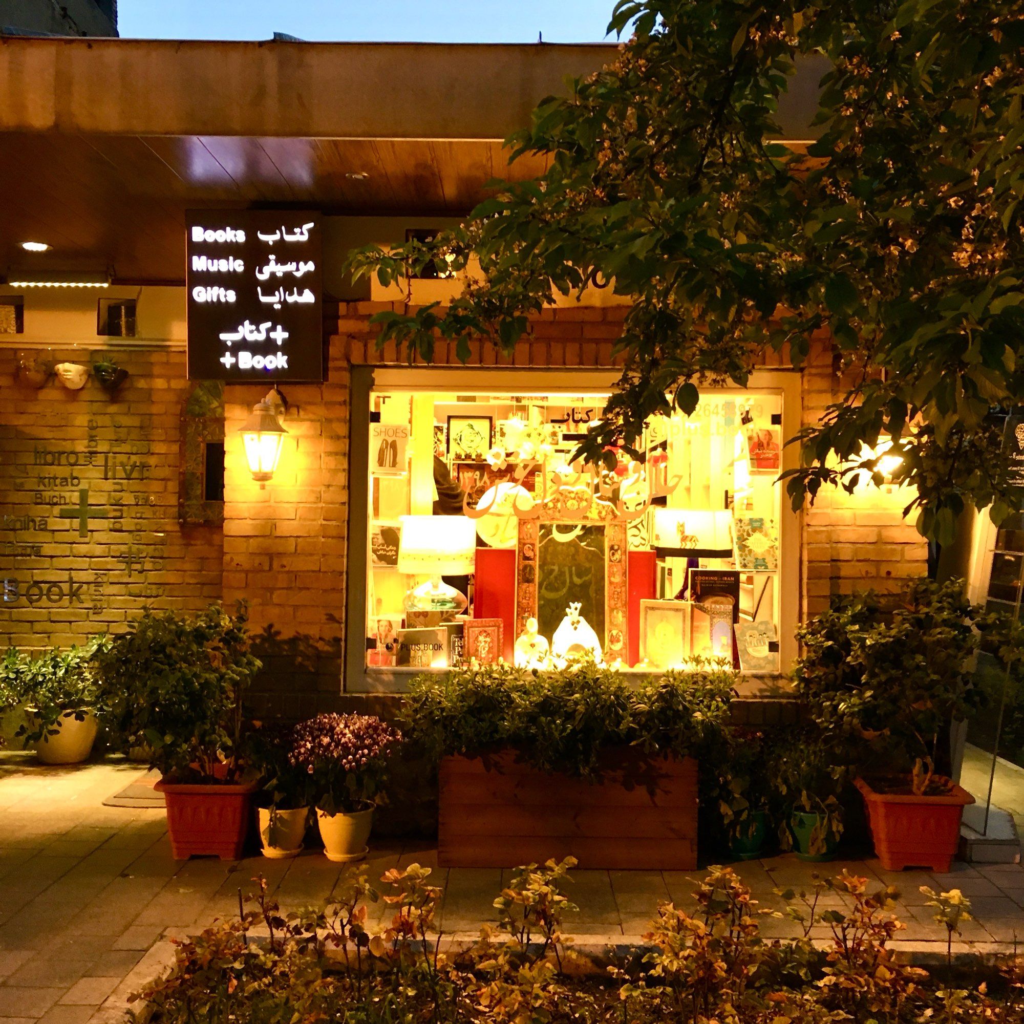 Store with lights on, brick wall, plants und flowers in pots; sign “books, music, gifts” in Persian and English