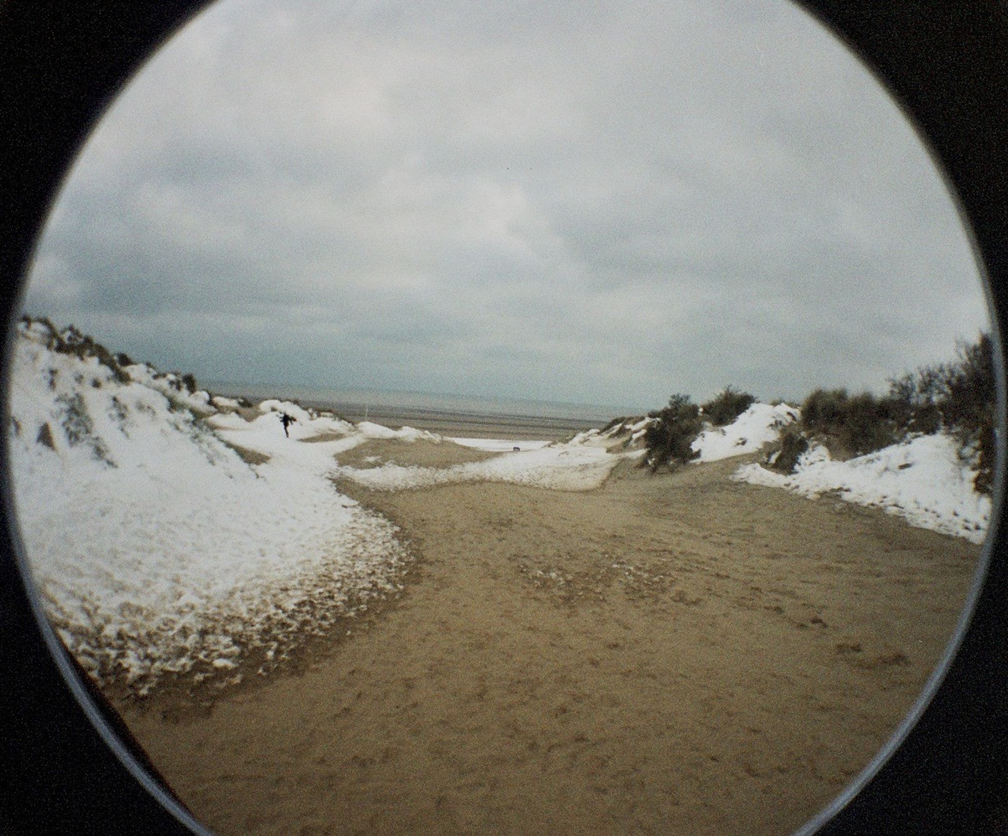 A fisheye photo of a sandy path to a beach, the dunes either side covered in snow