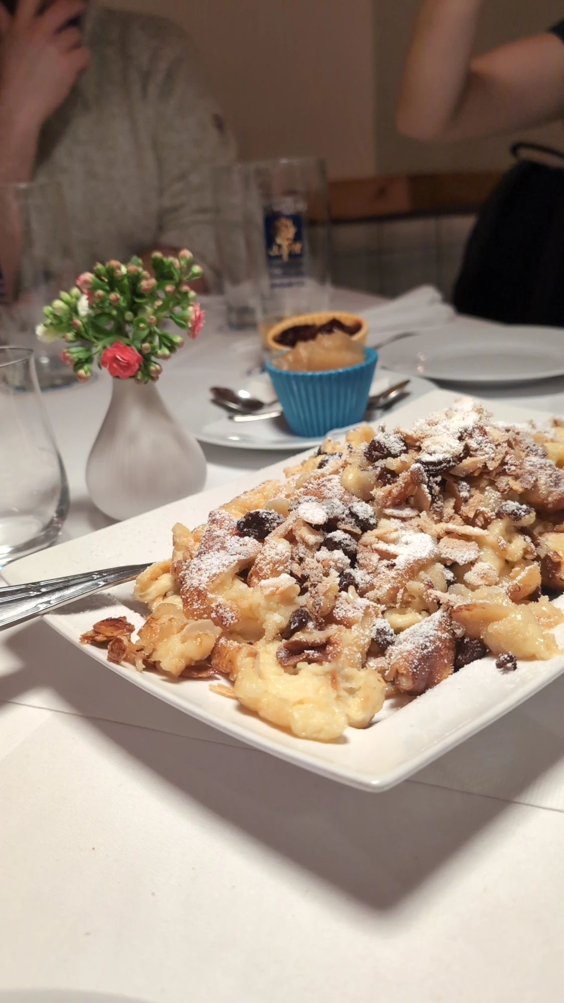 Ein großer eckiger Teller voll Kaiserschmarrn mit Rosinen, arrangiert neben einer kleinen Blumenvase und Muffinähnlicher Töpfchen mit Apfelkompott und Zwetschgenröster