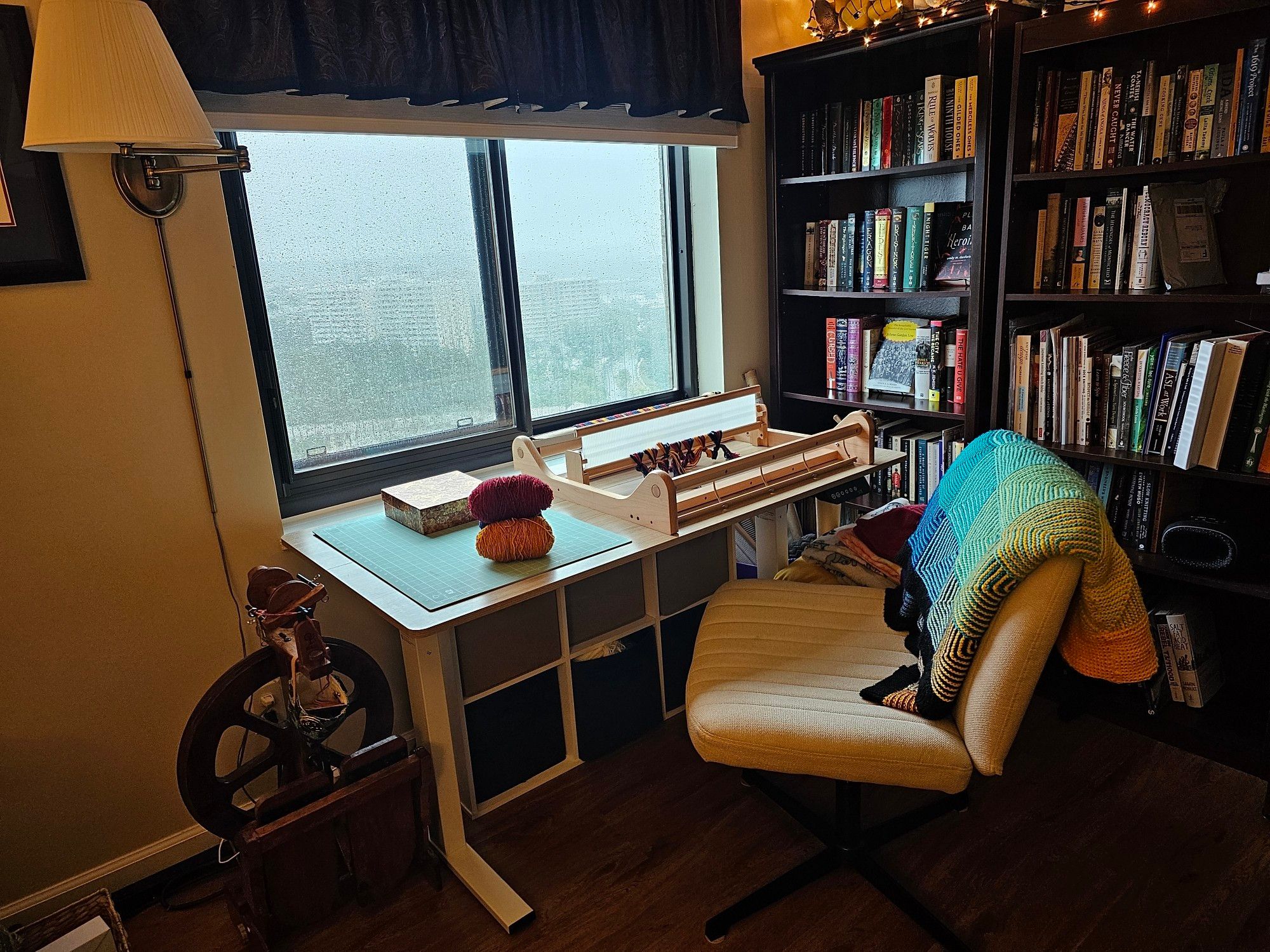 A desk with a rigid heddle loom, cutting mat, and yarn sits in front of a window on a rainy afternoon. To the left od the desk is a spinning wheel. To the right of the desk are bookcases with string lights running across the top. A rainbow blanket rests over the chair in front of the desk.