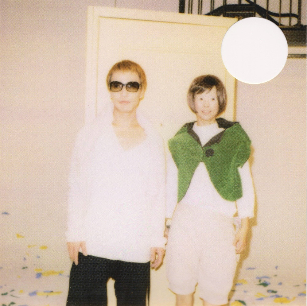 A photo of the members of capsule. Yasutaka Nakata wears a white long sleeve top, black trousers, and sunglasses. Toshiko Koshijima wears a white high-neck top, cream knee-length balloon pants, and a fuzzy green vest. They stand in front of a yellow door and a white backdrop, surrounded by confetti. 