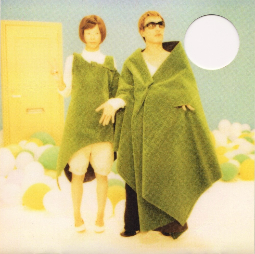 A photo of the members of capsule. Yasutaka Nakata wears a green robe, a white long sleeve, black trousers, and sunglasses. His hair is blond and cut short. Toshiko Koshijima wears a green asymmetrical dress of the same material over a white high neck top, cream knee-length balloon pants, and white ballet flats. Her brown hair is cut into a neat, round bob. They stand in a green room with a yellow door, surrounded by green, yellow, and white balloons. 