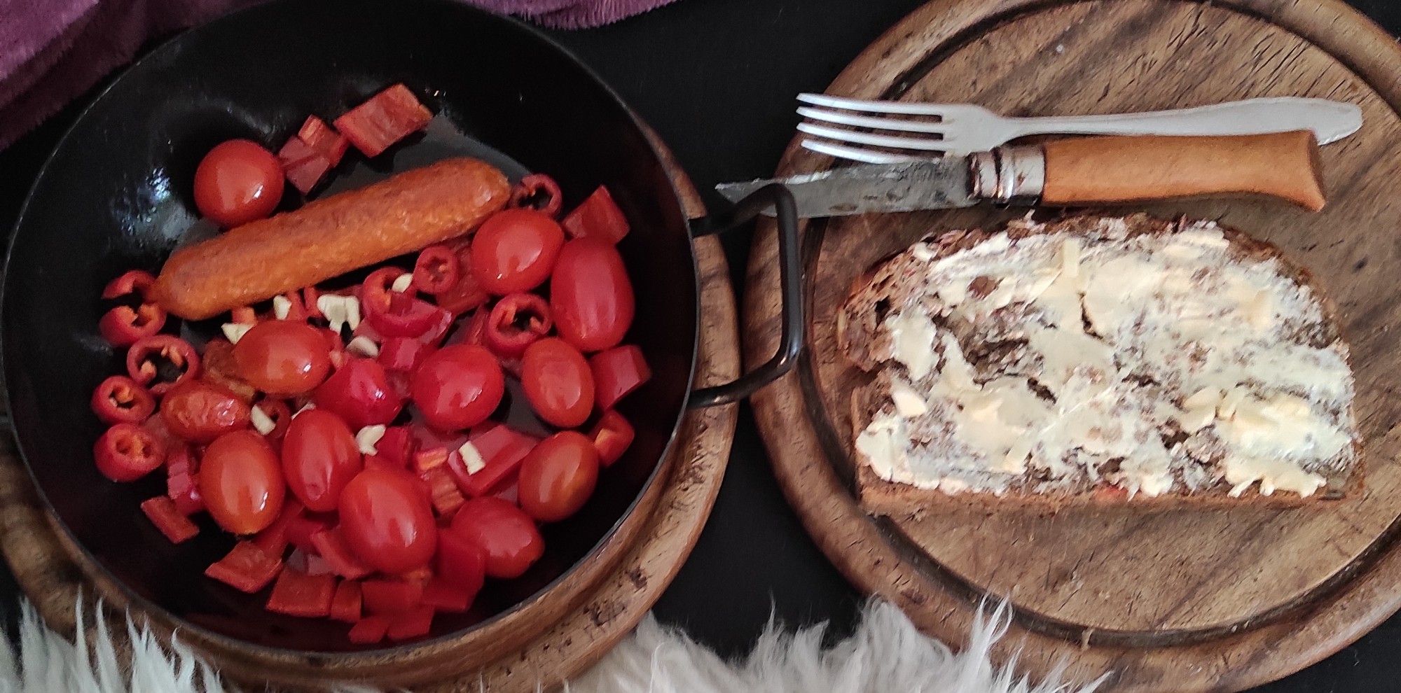 Kleine Bratpfanne mit Bratwurst, Cherrytomaten, roter Spitzpaprika (kleingeschnitten), roter Peperoni (kleingeschnitten) und Knoblauch (kleingeschnitten) auf Holzbrett.

Daneben Holzbrett mit Stulle mit veganer Butter und Knoblauch (kleingeschnitten) und Gabel mit Opinelmesser.