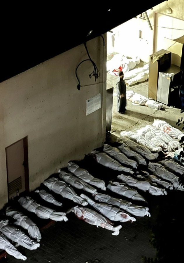 A darkened street next to a white wall and floor is covered in bodies lined up on the floor in their white shrouds.