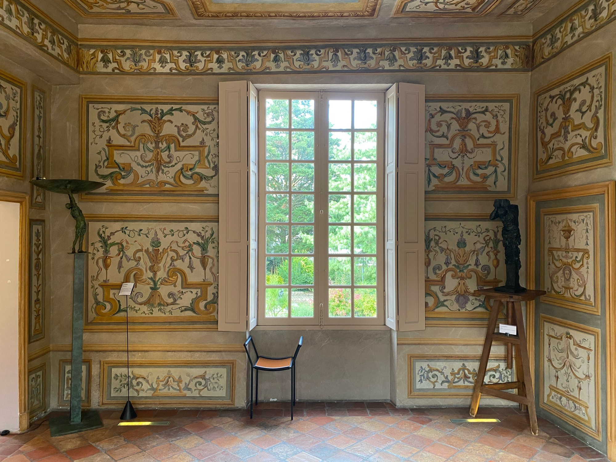 Photo d’une salle quasiment vide avec une large baie vitrée. Au mur sont peints des décors d’arabesques et de motifs végétaux du XVIIe siècle. 