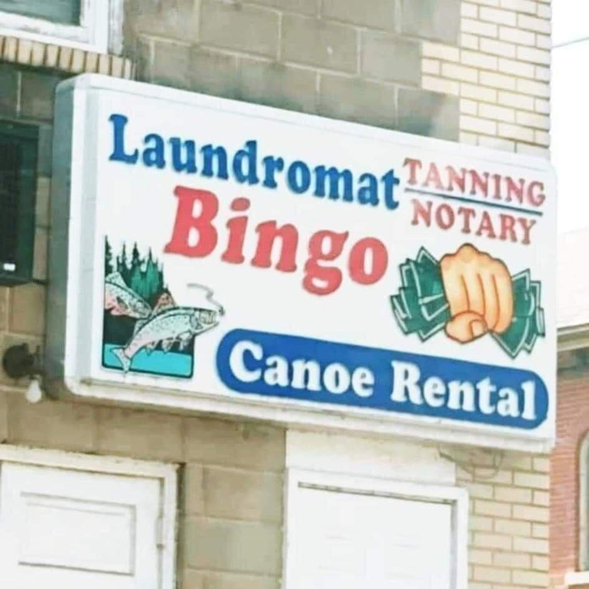 A business sign that reads Laundromat, Tanning / Notary, Bingo, and Canoe Rental, with Tanning and Notary above and below a line, a picture of a fist clenching money, and a fish leaping out of the water.